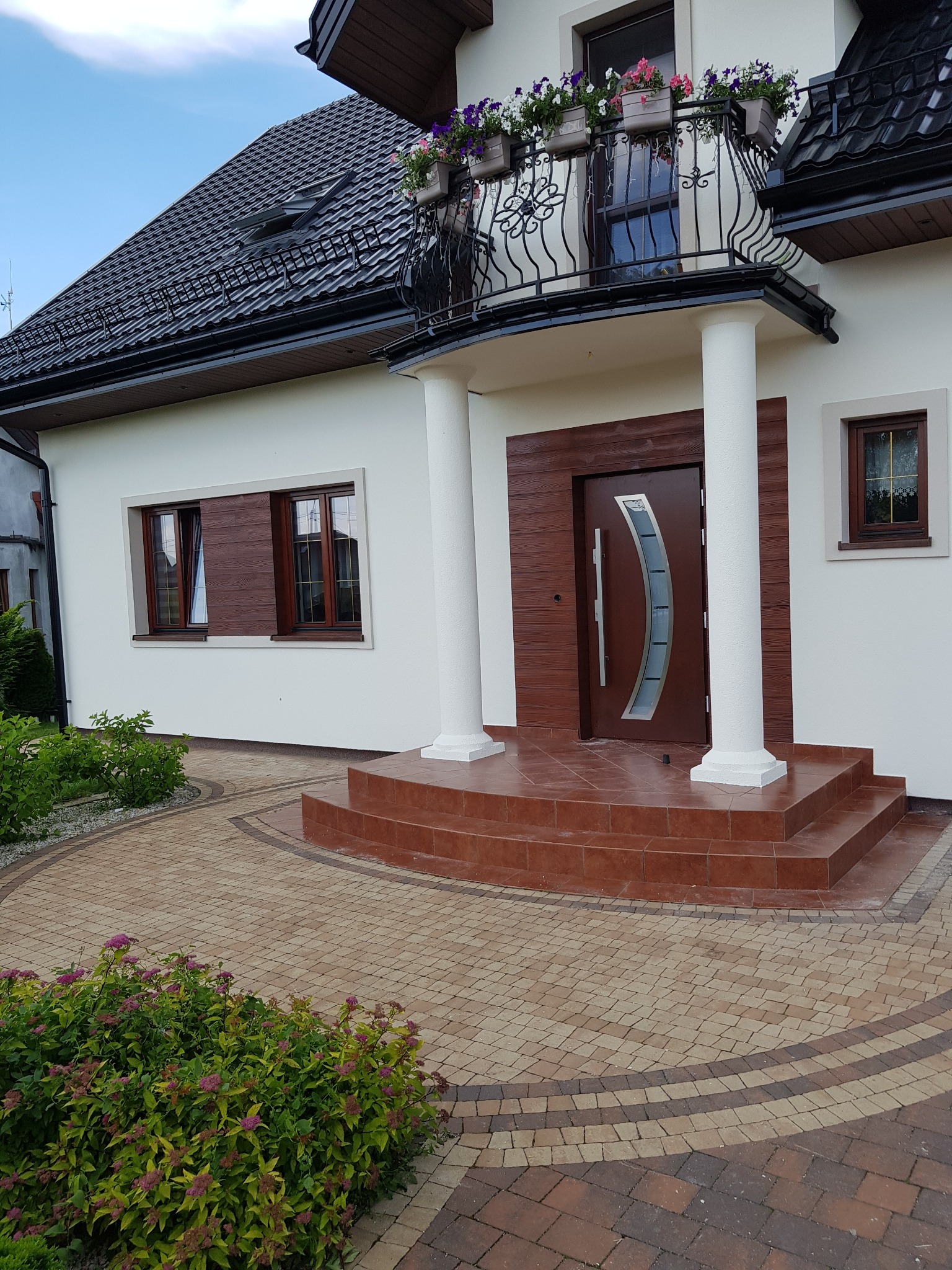 Biała elewacja domu jednorodzinnego z brązowymi oknami i drzwiami, kolumnami przy wejściu oraz brukowanym podjazdem z roślinnością.