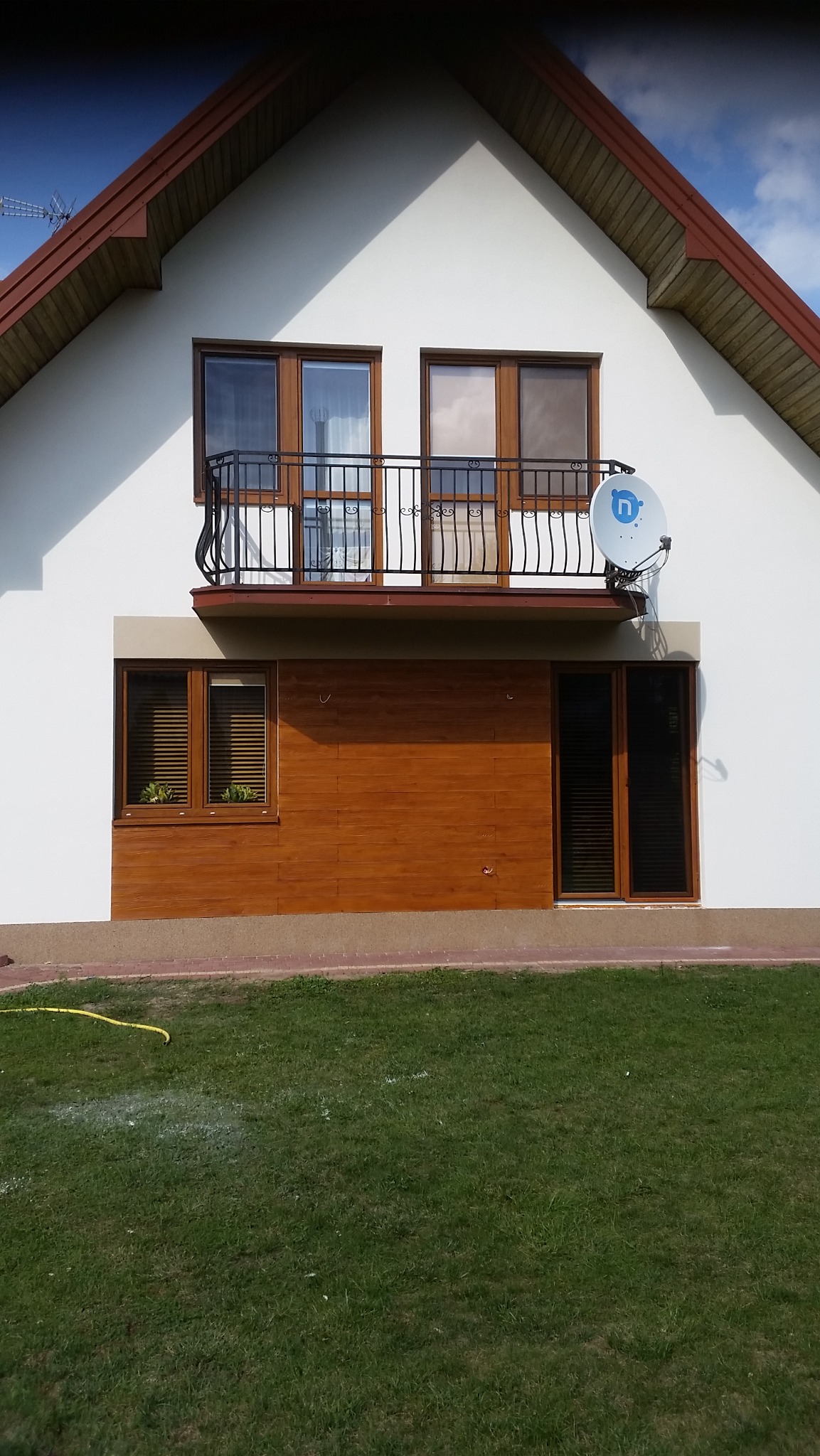 Ocieplony dom jednorodzinny z białymi ścianami, brązowymi oknami i balkonem z czarną balustradą, widoczny satelitarny talerz antenowy.