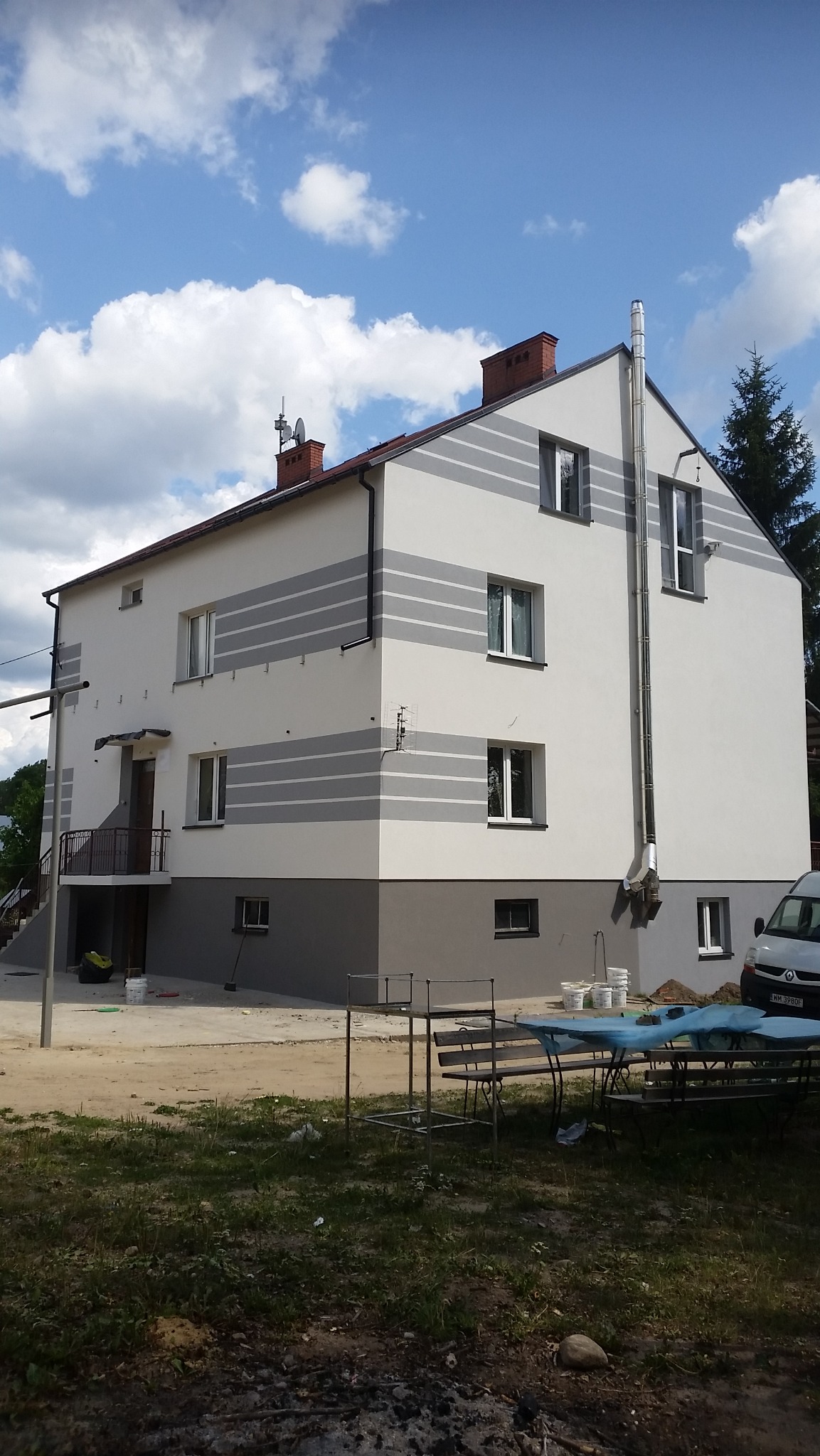 Dwukondygnacyjny dom po remoncie elewacji, z szarymi pasami na białej fasadzie, widoczne ślady prac wykończeniowych, w tle błękitne niebo z chmurami.