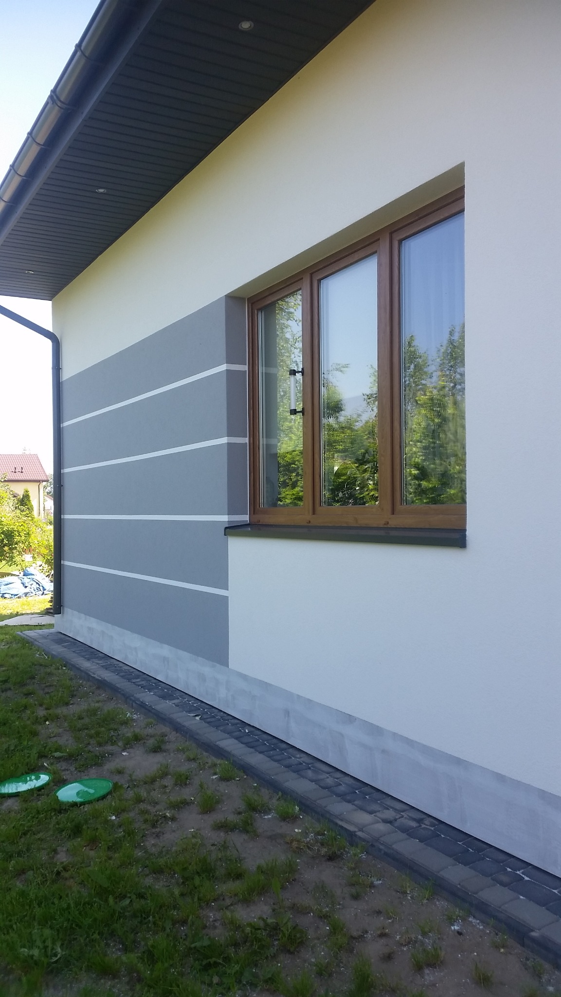 Fragment ściany domu z oknem, szara elewacja z poziomymi białymi paskami, widoczne zielone pokrywy studzienek na trawniku.