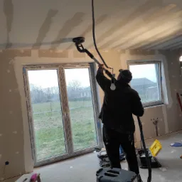 Pracownik w czapce szlifuje sufit szlifierką teleskopową z odkurzaczem w pomieszczeniu z dużymi oknami, widoczne ślady po łączeniach płyt gipsowych.