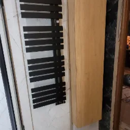 Nowoczesna łazienka z czarnym grzejnikiem drabinkowym na ścianie z marmurowym wzorem, obok drewniana szafka wisząca i częściowo widoczna kabina prysznicowa z czarnymi profilami.