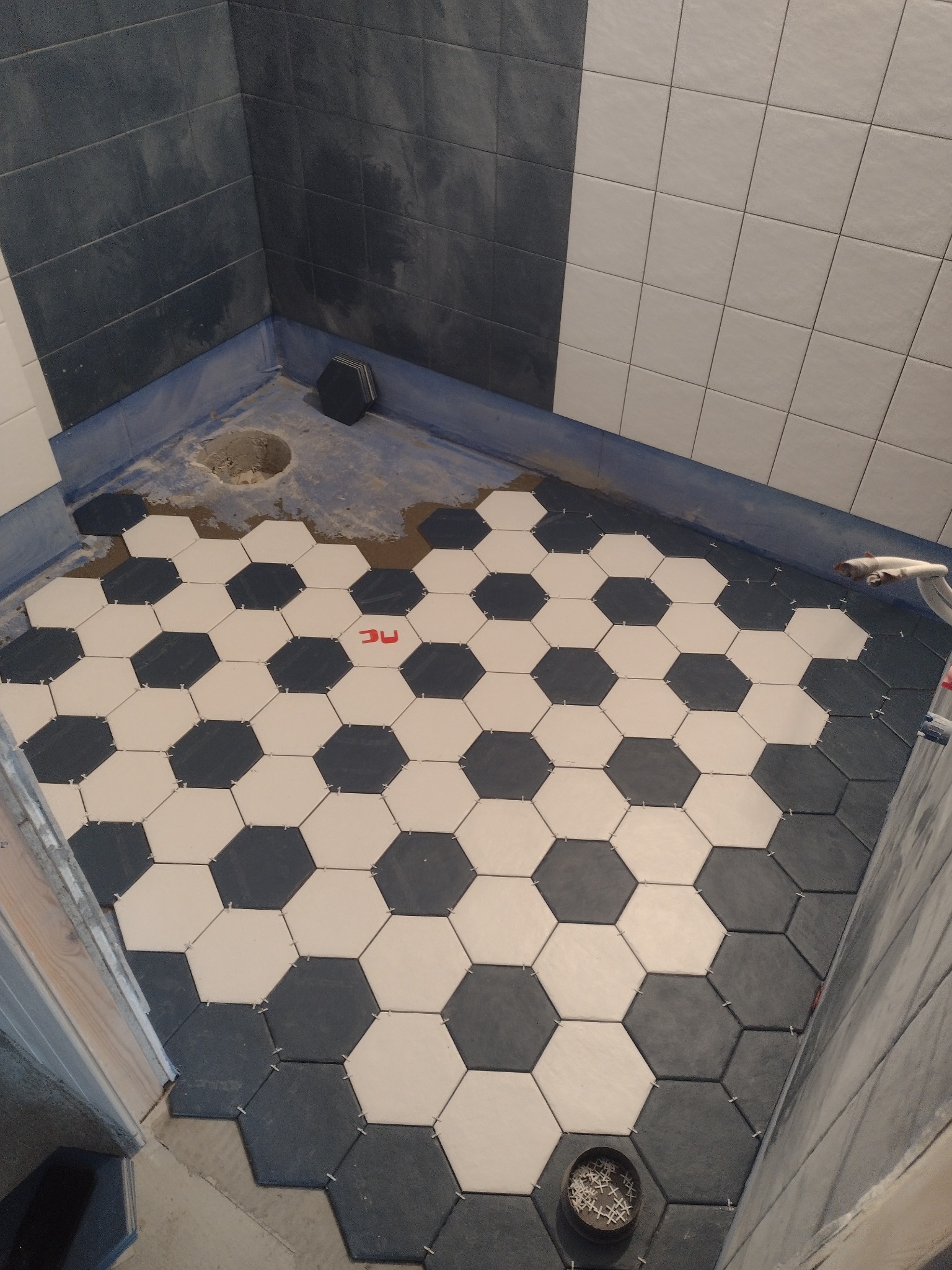 Układanie heksagonalnych płytek w kolorze białym i grafitowym na podłodze prysznica; widoczne krzyżyki dystansowe, odpływ oraz fragment ściany z szarymi i białymi płytkami.