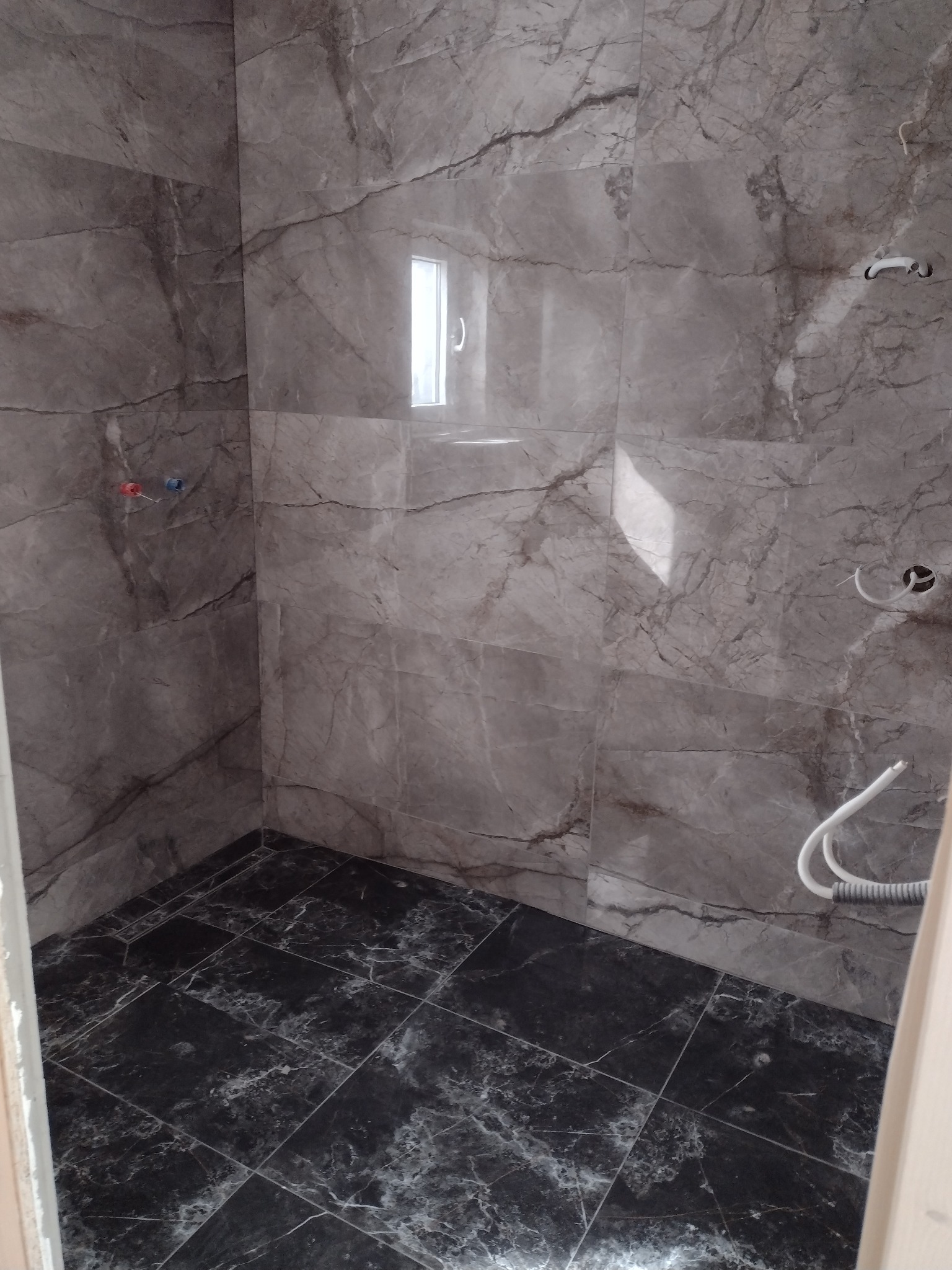 Wnętrze łazienki w trakcie prac wykończeniowych, widoczne szare marmurowe płytki na ścianach i czarne na podłodze, wystające rury instalacji wodnej i elektrycznej, małe okno w górnej części ściany.