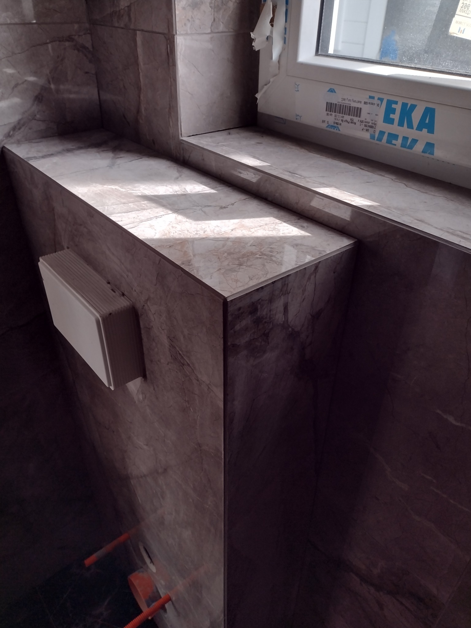 Wykończona wnęka okienna i fragment ściany w łazience, obłożone płytkami imitującymi marmur. Widoczne podejście instalacji wodnej i biała obudowa przyłącza.