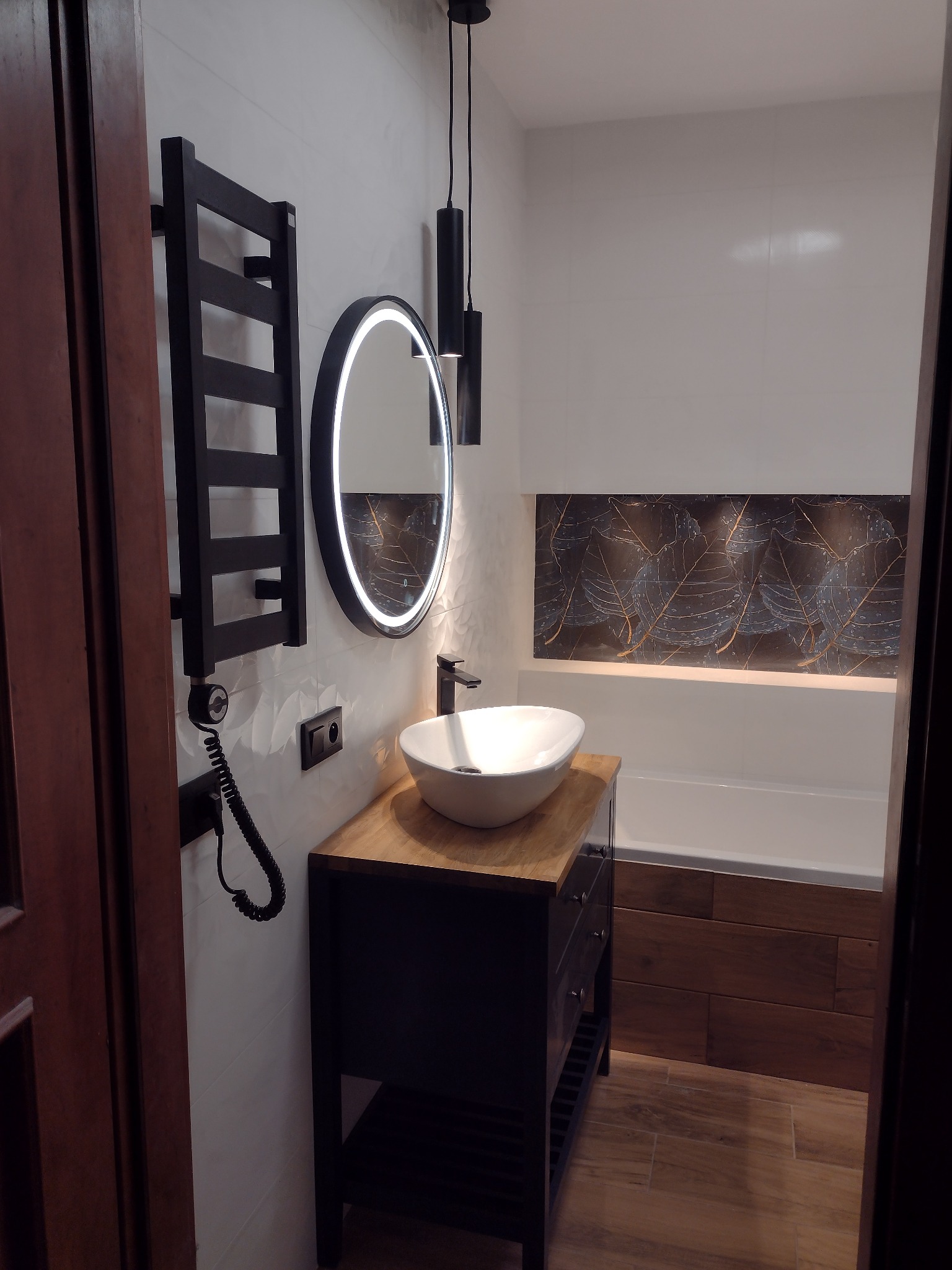 Nowoczesna łazienka z umywalką nablatową, podświetlanym lustrem, czarnym grzejnikiem drabinkowym i płytkami z motywem liści w strefie wanny.