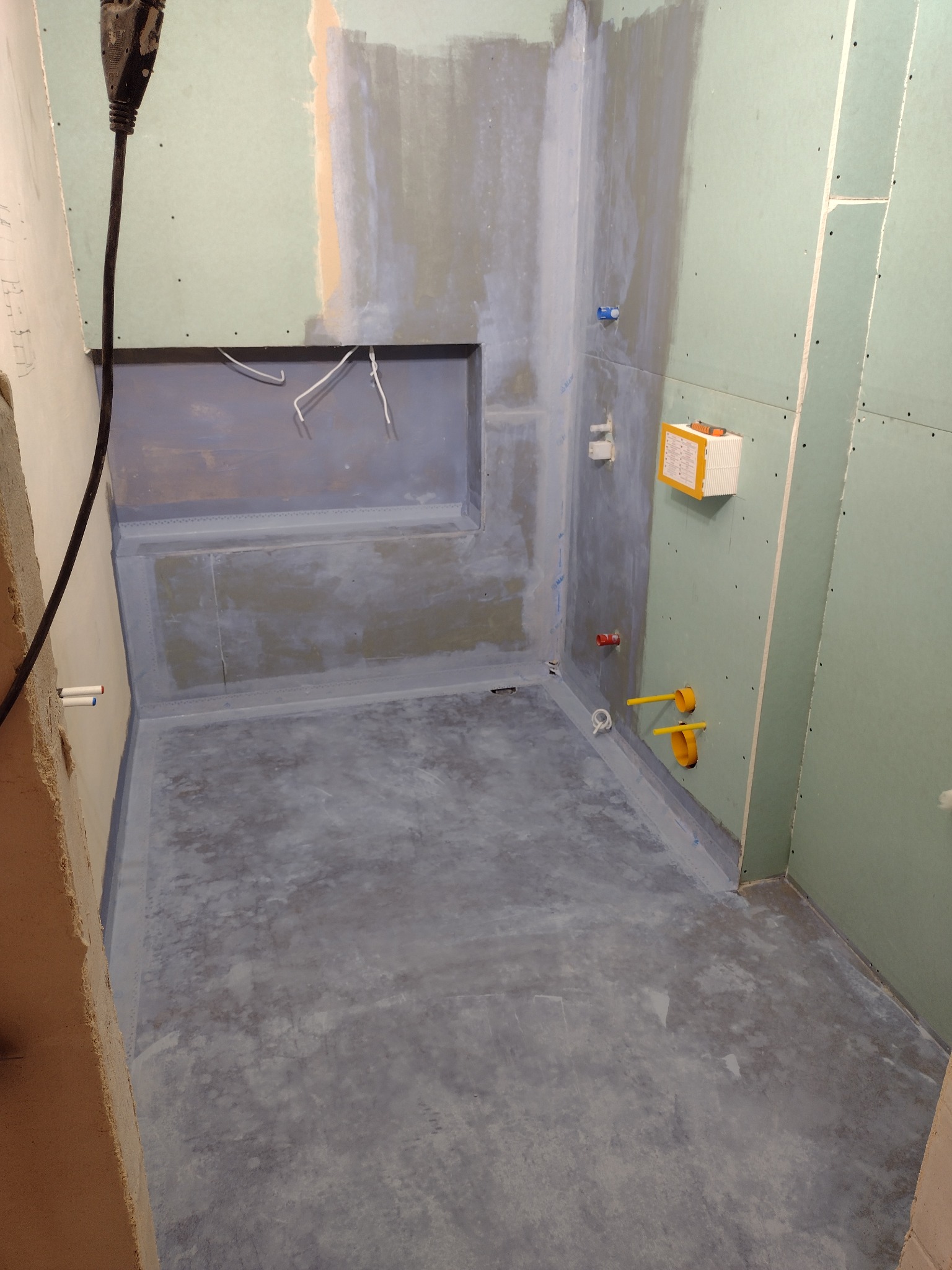 Przygotowana do hydroizolacji łazienka w trakcie prac: zagruntowana podłoga i ściany, widoczne fragmenty płyt karton gips, wystające rury instalacji wodnej i elektrycznej, oraz kabel zasilający...