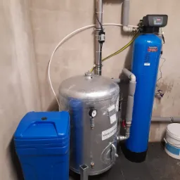 Instalacja hydroforowa: srebrny zbiornik hydroforowy z manometrem, niebieski zmiękczacz wody z elektronicznym panelem sterowania oraz niebieski zasobnik na sól, rury ocieplone szarą pianką...