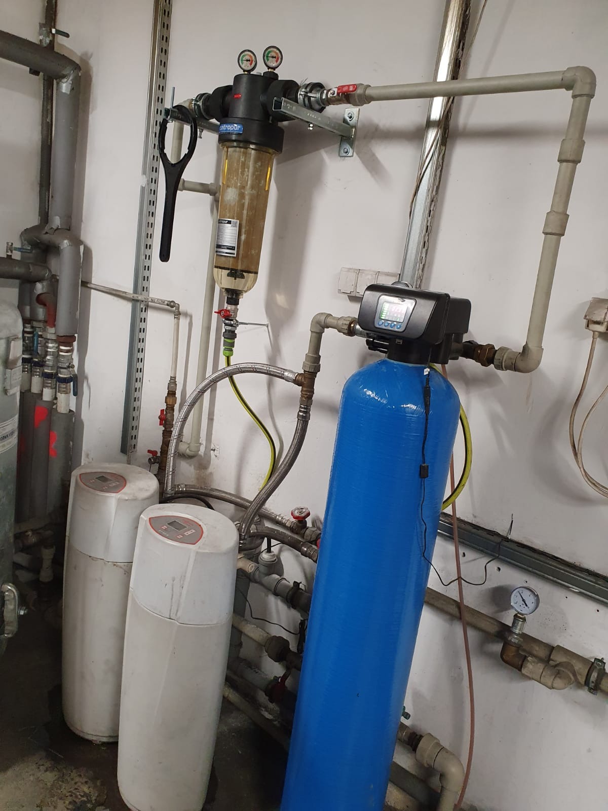 System filtracji wody z kolumną niebieską i filtrem mechanicznym z brązowym osadem, manometry, rury, elementy instalacji w pomieszczeniu technicznym.