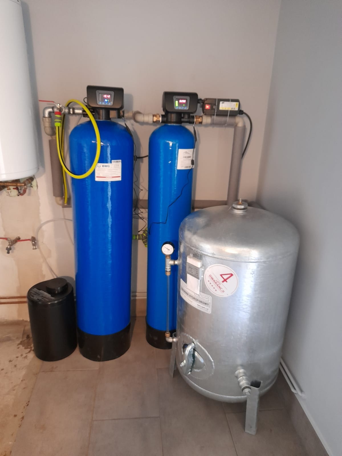 Dwa niebieskie filtry kolumnowe z elektronicznymi wyświetlaczami, obok srebrny zbiornik hydroforowy z naklejką '4 lata gwarancji', zainstalowane w pomieszczeniu z szarą ścianą i beżową podłogą.