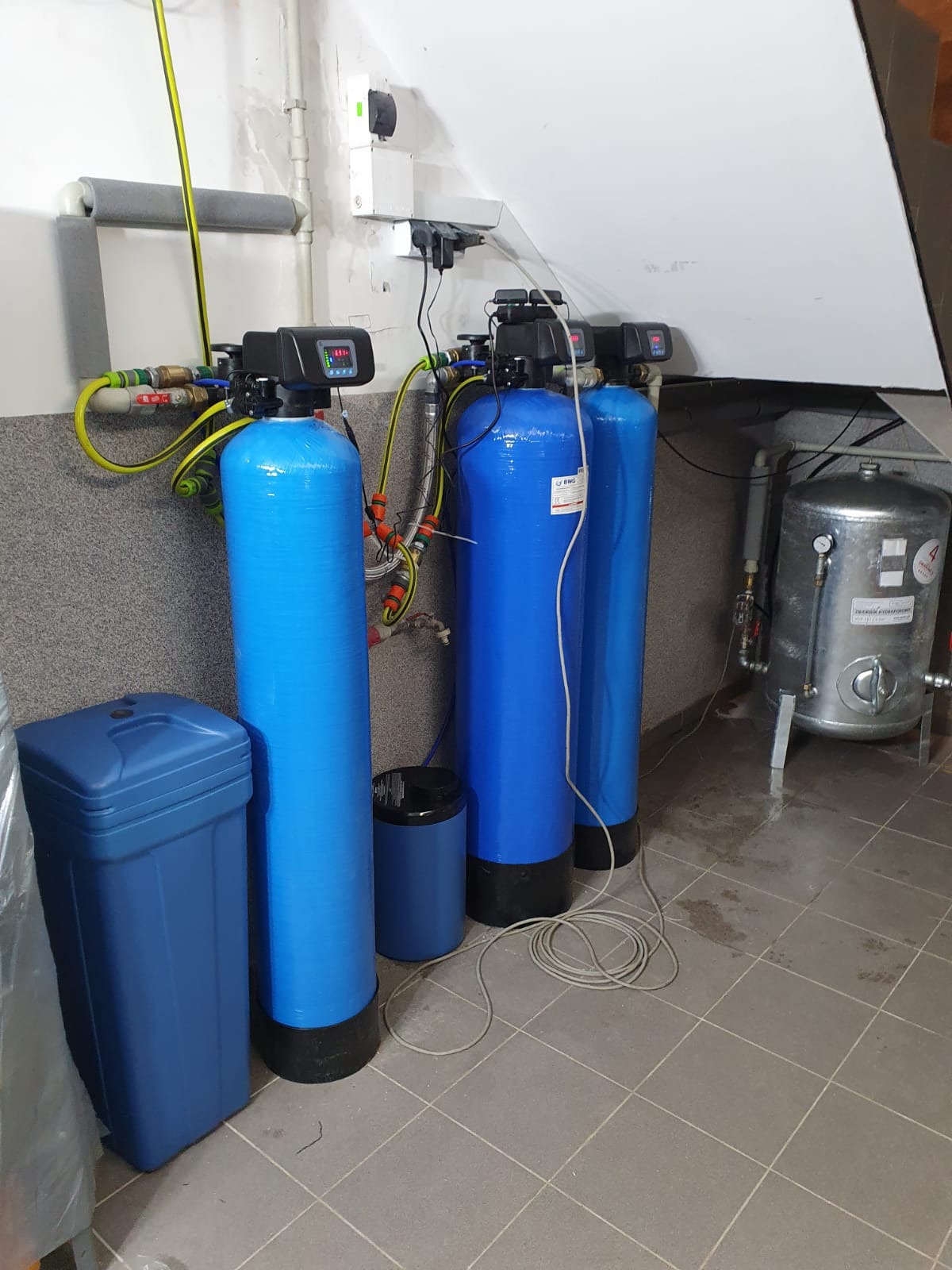 Instalacja filtrująca wodę w piwnicy, składająca się z trzech niebieskich cylindrycznych zbiorników z czarnymi głowicami, połączonych rurami i przewodami, obok zbiornik ciśnieniowy, na podłodze...