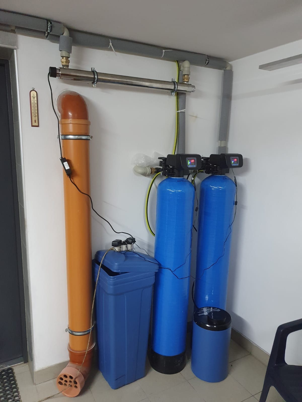 Domowa instalacja uzdatniania wody: dwa niebieskie filtry ciśnieniowe z wyświetlaczami, zbiornik na sól, rura odpływowa i lampa UV zamontowana pod sufitem w pomieszczeniu technicznym.