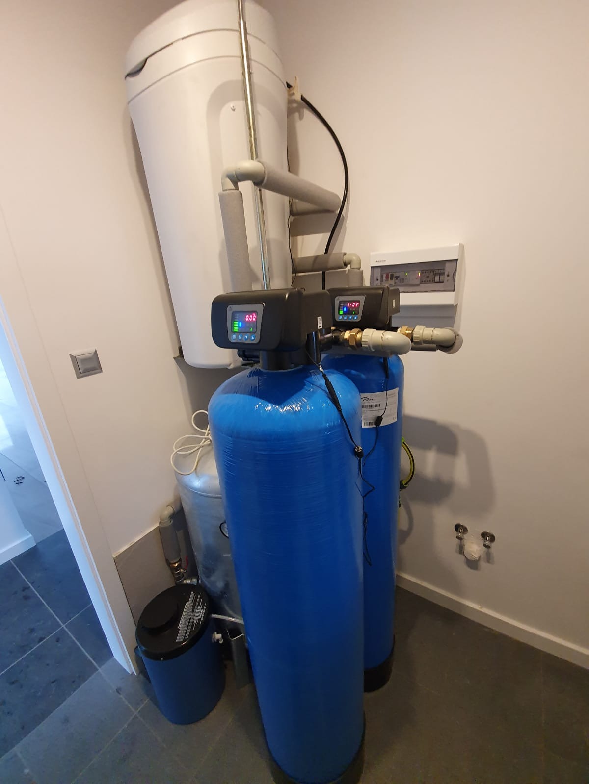 Dwa niebieskie zbiorniki z czarnymi głowicami sterującymi, podłączone rurami, obok biały zbiornik ciśnieniowy i skrzynka elektryczna na ścianie w pomieszczeniu z szarą podłogą.