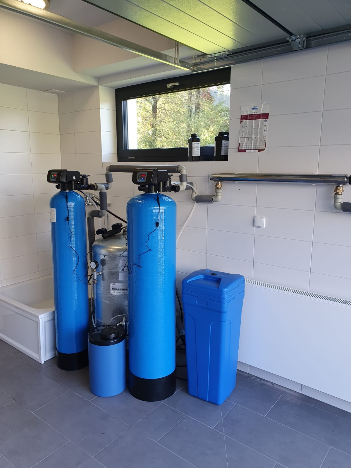 Instalacja filtrów wody w pomieszczeniu z oknem, widoczne dwa niebieskie filtry kolumnowe z elektronicznymi wyświetlaczami, zbiornik hydroforowy, niebieski zasobnik na sól, białe płytki na ścianach...