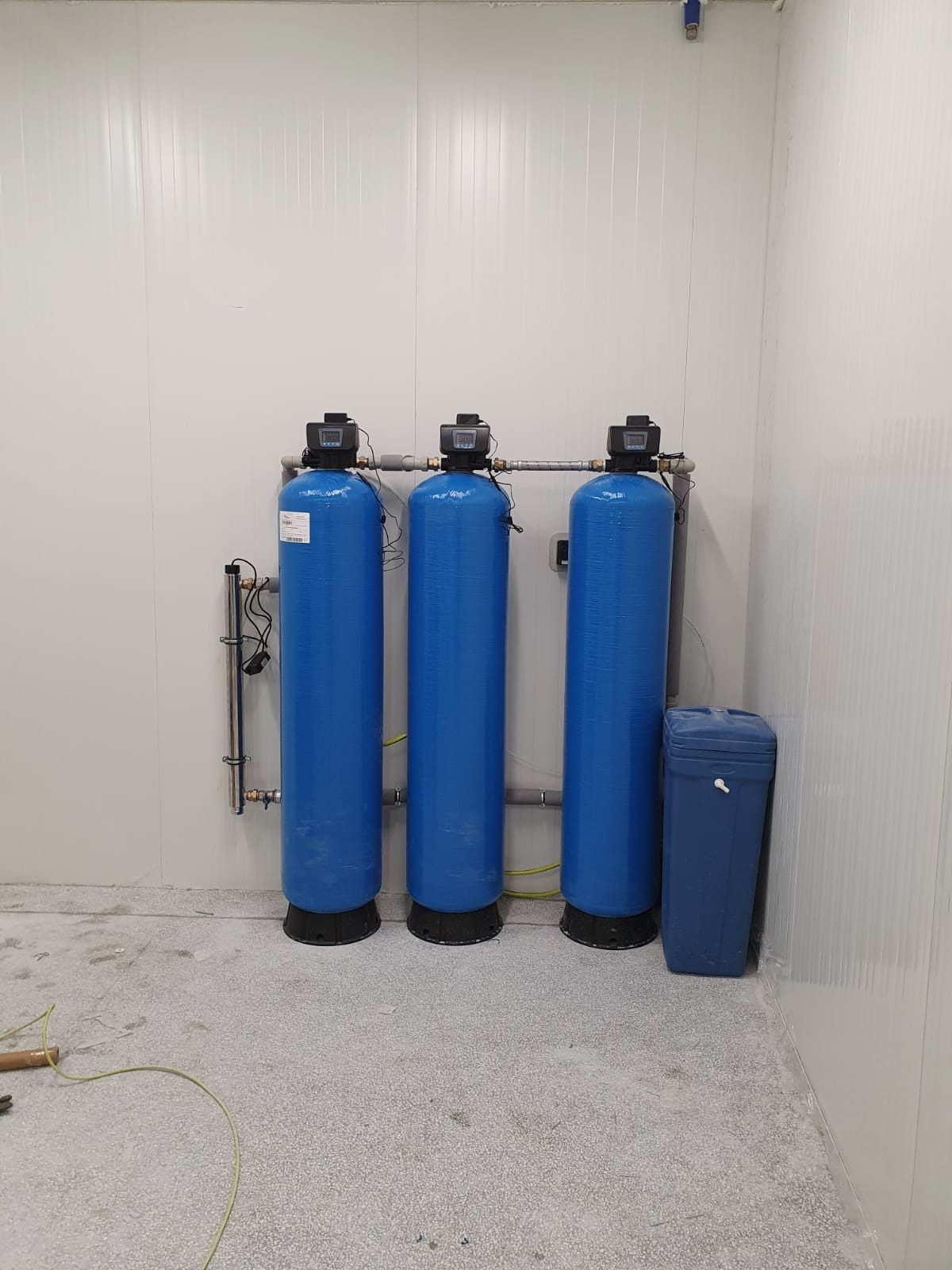 Trzy niebieskie, pionowe zbiorniki ciśnieniowe z czarnymi głowicami, połączone szarymi rurami, stojące przy ścianie z paneli w sterylnym pomieszczeniu. Obok skrajnego prawego zbiornika stoi...