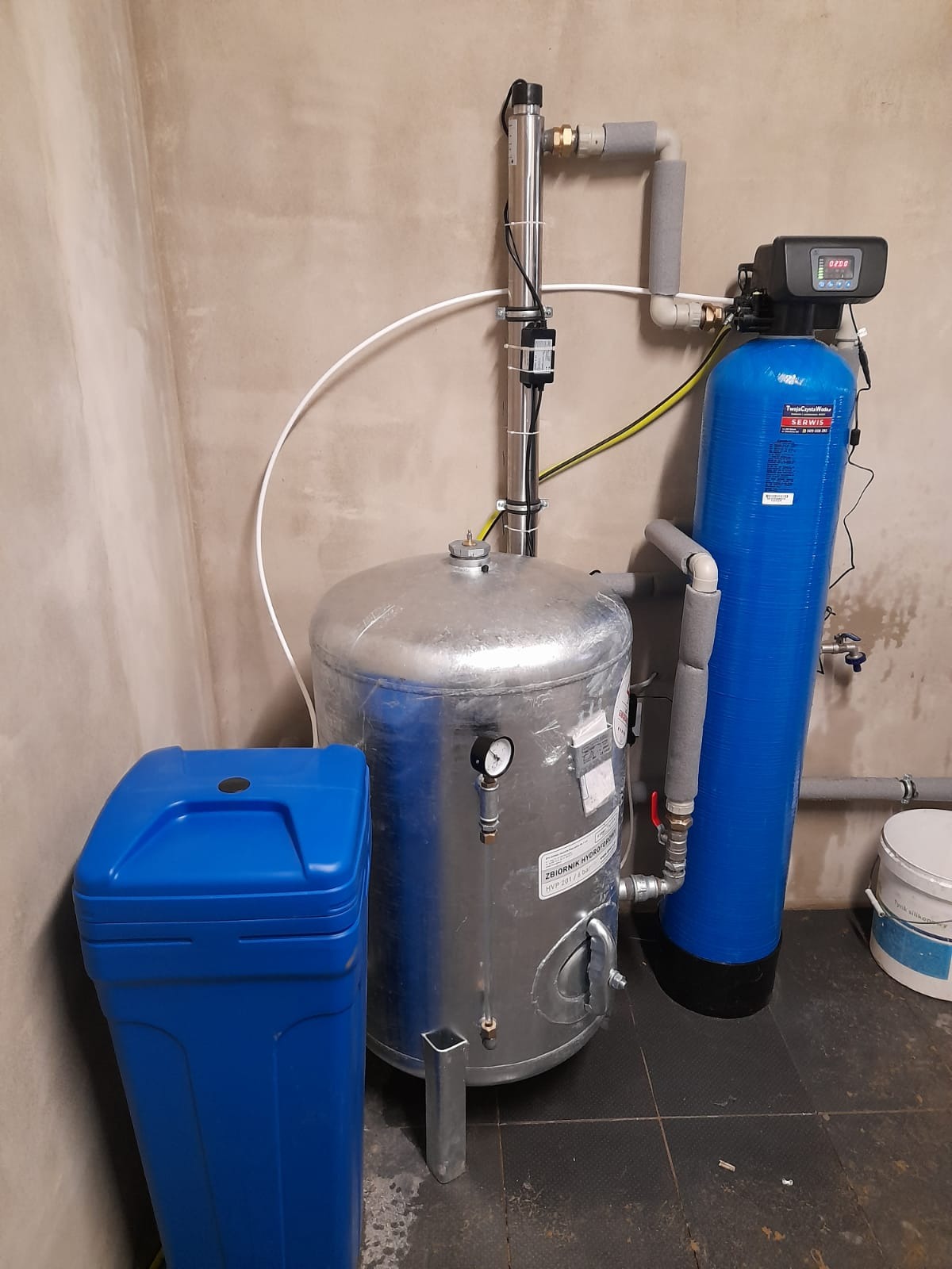 Instalacja hydroforowa: srebrny zbiornik hydroforowy z manometrem, niebieski zmiękczacz wody z elektronicznym panelem sterowania oraz niebieski zasobnik na sól, rury ocieplone szarą pianką...