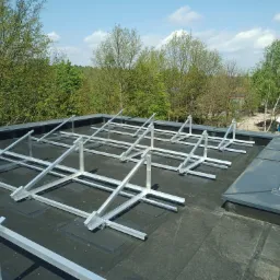 Konstrukcja wsporcza pod panele fotowoltaiczne na czarnym, płaskim dachu. Widok z góry, w tle zieleń drzew i błękitne niebo. System balastowy i klejony.