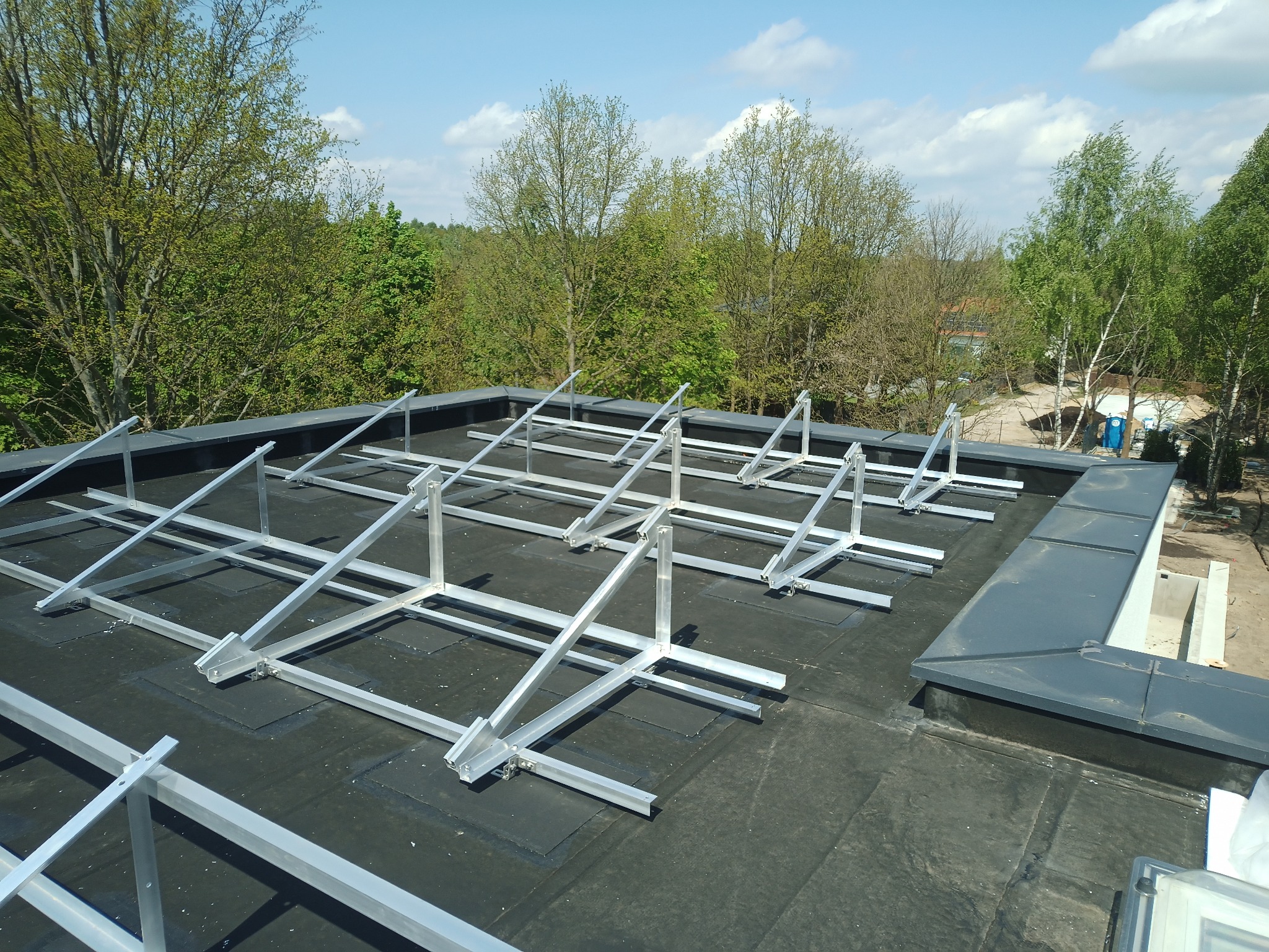 Konstrukcja wsporcza pod panele fotowoltaiczne na czarnym, płaskim dachu. Widok z góry, w tle zieleń drzew i błękitne niebo. System balastowy i klejony.