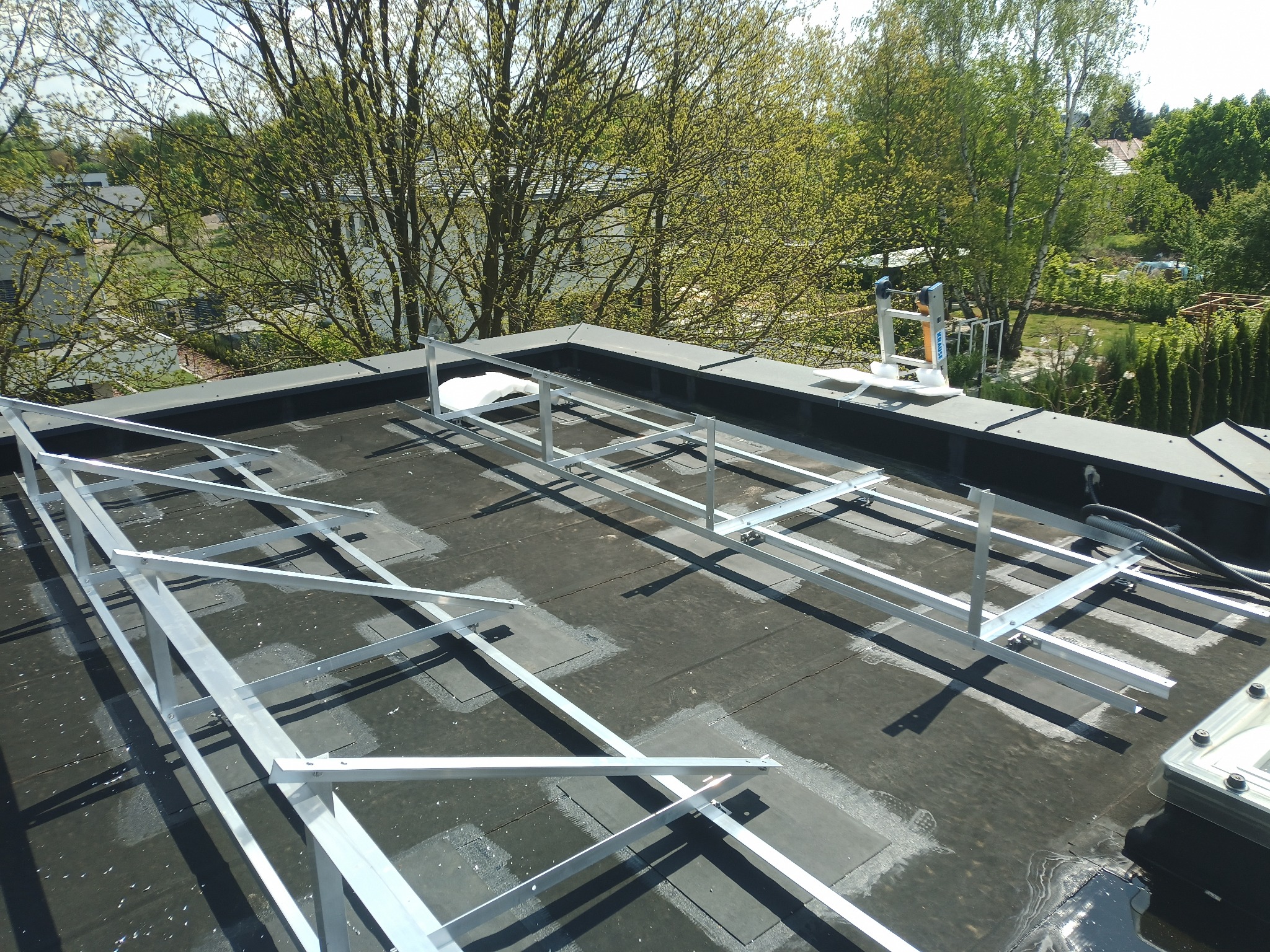 Montaż konstrukcji wsporczej pod panele fotowoltaiczne na płaskim dachu pokrytym papą. Widoczne elementy mocujące i przewody. Tło stanowi zieleń drzew i zabudowa.