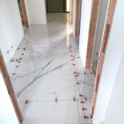 Rafał lewicki - Układanie białych płytek podłogowych z szarym wzorem marmuru w korytarzu, widoczne czerwone klipsy poziomujące i niedokończone ościeżnice drzwi.