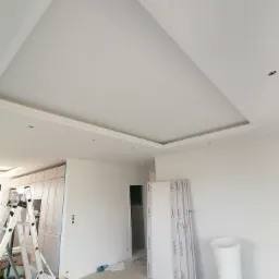 Biały sufit z podwieszanym fragmentem i dekoracyjną listwą, przygotowany pod montaż oświetlenia, widoczne otwory, w tle drabina i przygotowane panele ścienne.