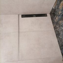 Rafał lewicki - Jasne, kwadratowe płytki podłogowe z czarnym odpływem liniowym, widoczne fragmenty marmurowej ściany i but roboczy w rogu kadru.