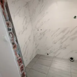 Wnętrze łazienki w trakcie remontu, widoczne płytki imitujące marmur na ścianach i podłodze, odsłonięta cegła przy ścianie, przygotowane otwory na armaturę.