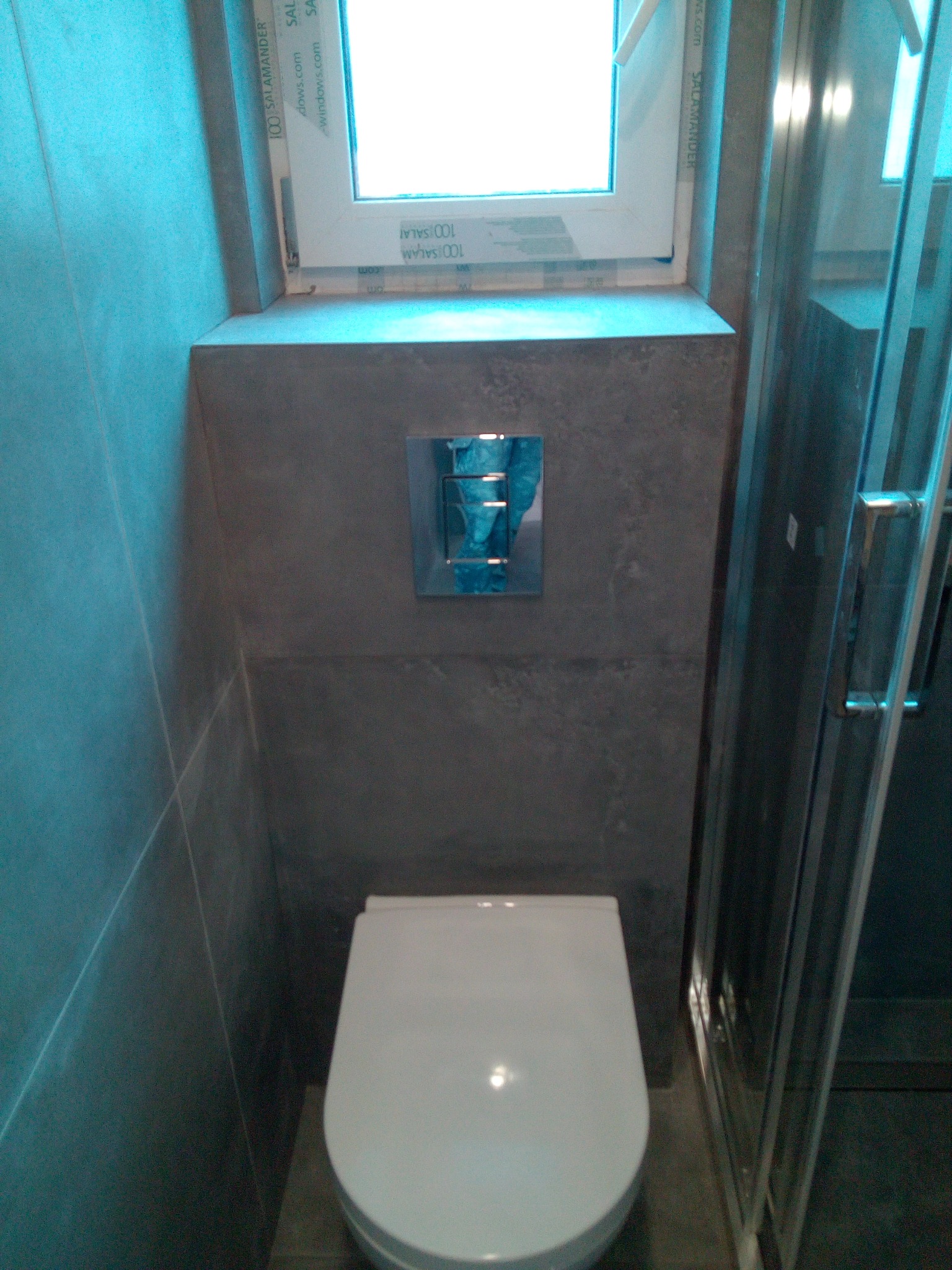 Nowoczesna toaleta wisząca w łazience wykończonej szarymi płytkami, z widocznym przyciskiem spłukującym i kabiną prysznicową w tle.