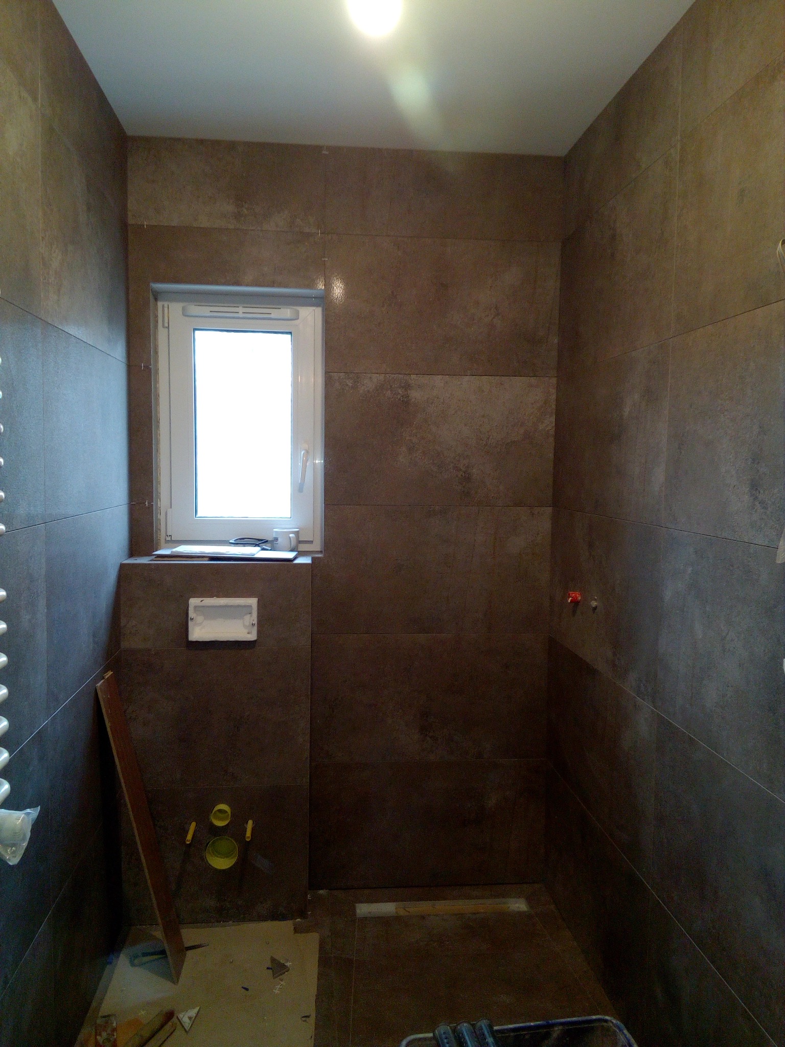 Łazienka w trakcie wykańczania: brązowe płytki na ścianach, okno z parapetem, widoczne elementy instalacji podtynkowych, narzędzia i materiały budowlane na podłodze.