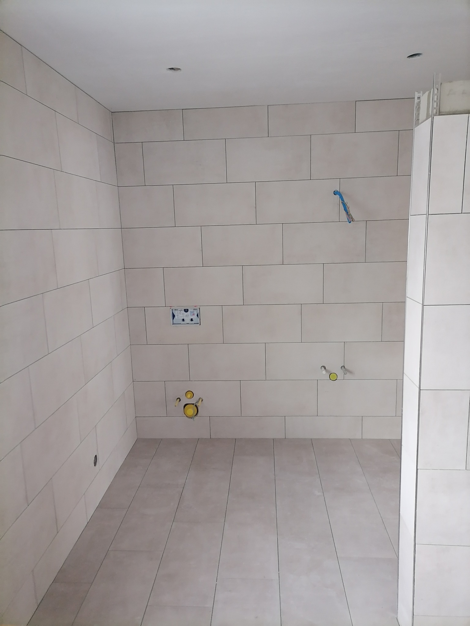 Wnętrze łazienki w trakcie prac wykończeniowych, widoczne ułożone jasne płytki ceramiczne na ścianach i podłodze, przygotowane instalacje wodno-kanalizacyjne z żółtymi zaślepkami i niebieskim...