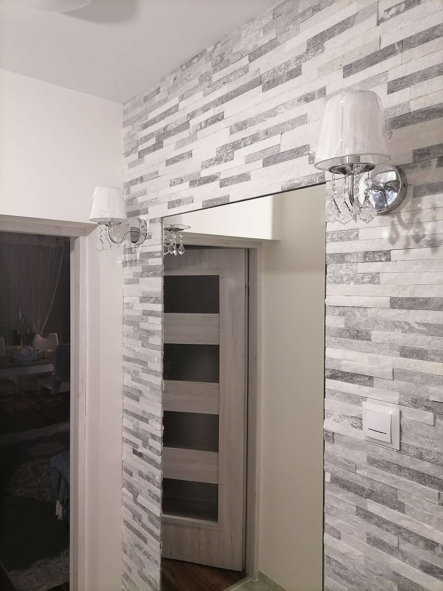 Ściana wykończona dekoracyjną płytką imitującą kamień w odcieniach szarości i bieli, oświetlona dwoma kinkietami z abażurami i kryształkami, widok na otwarte drzwi z płycinami.