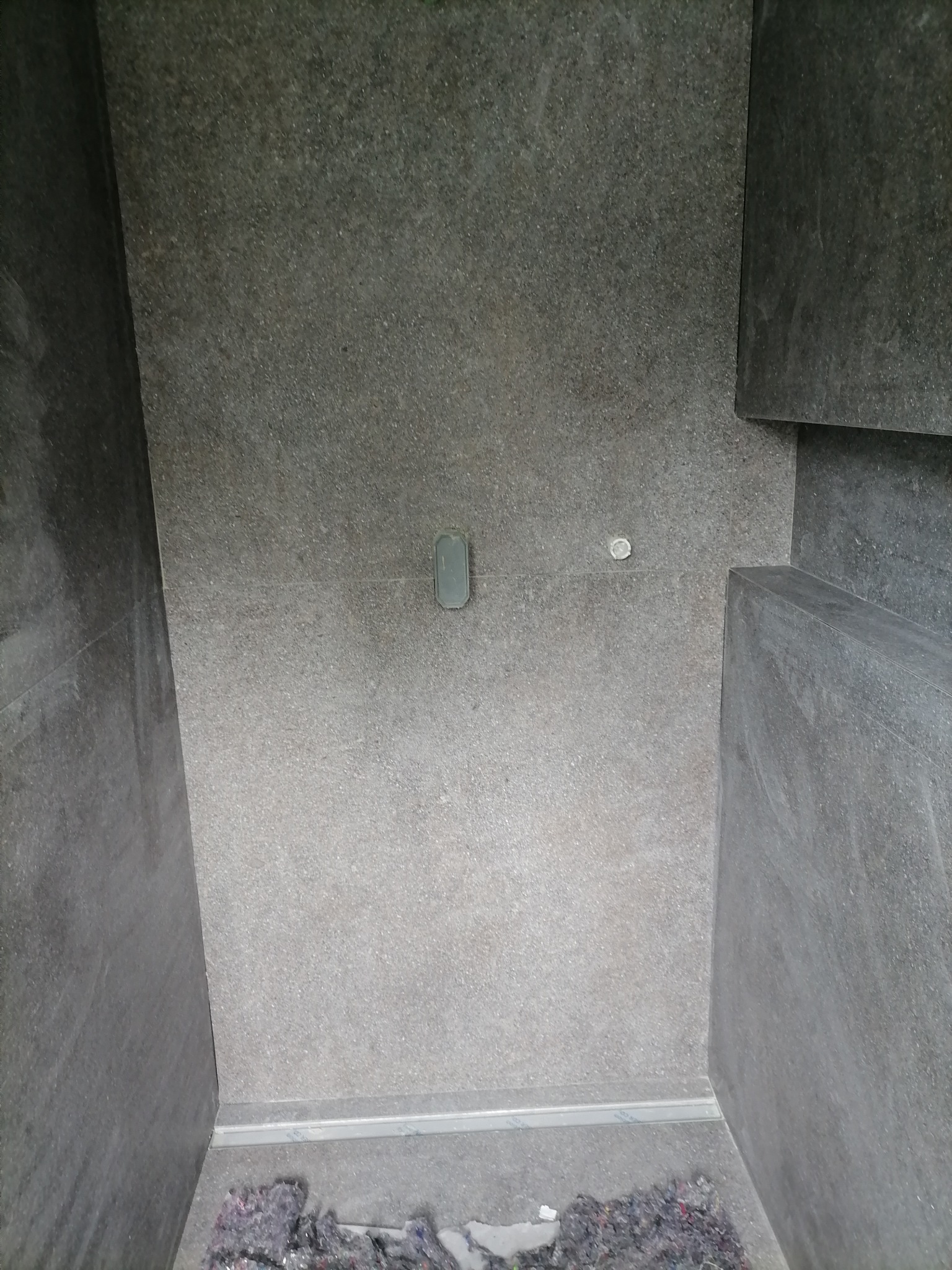 Fragment łazienki z szarą, matową okładziną ścienną i podłogową; widoczny odpływ liniowy i resztki materiałów budowlanych na podłodze.