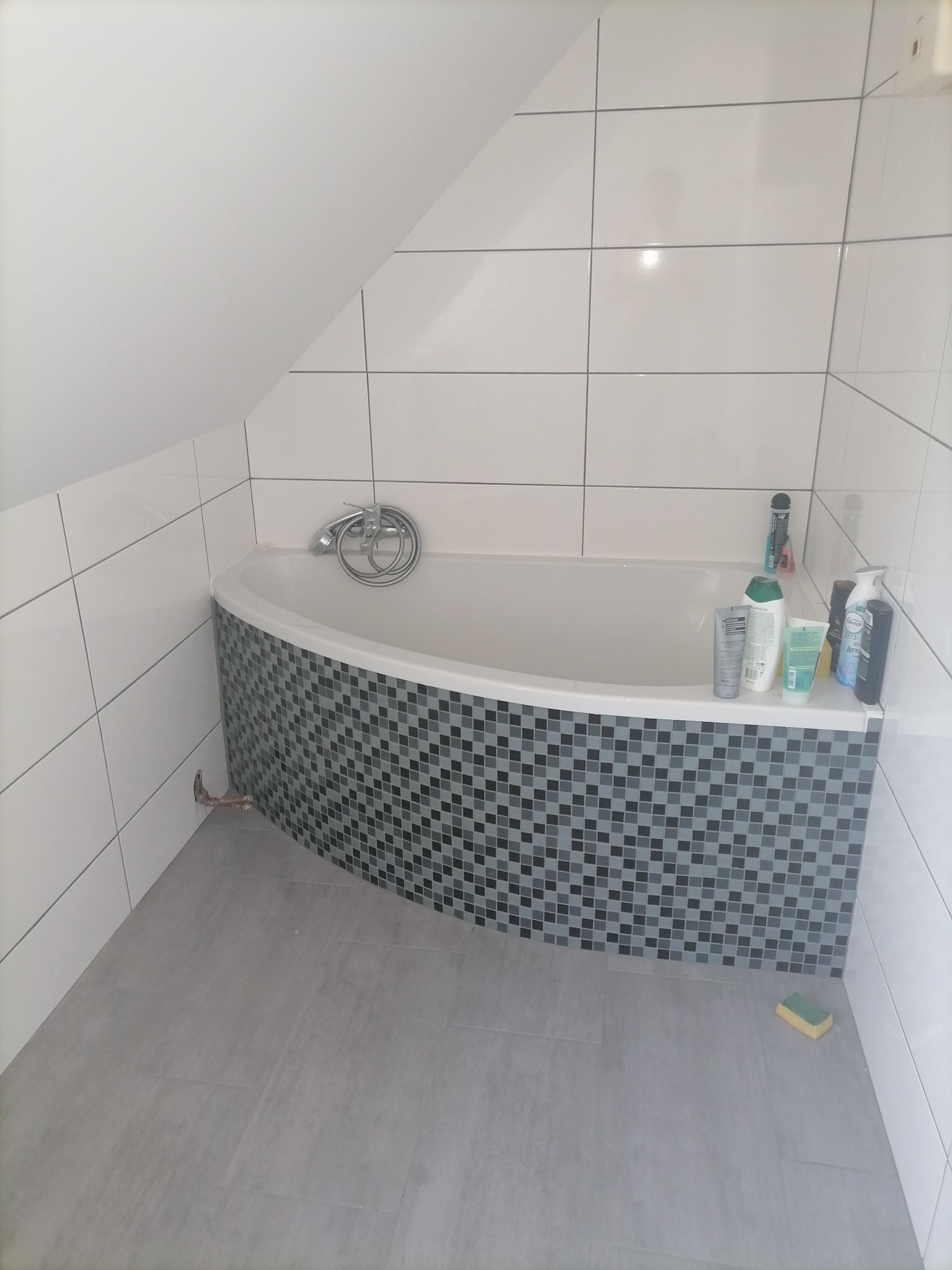 Narożna wanna obłożona mozaiką w odcieniach szarości i czerni, białe płytki na ścianach, szara podłoga, kosmetyki na krawędzi wanny, widoczna rura od baterii.