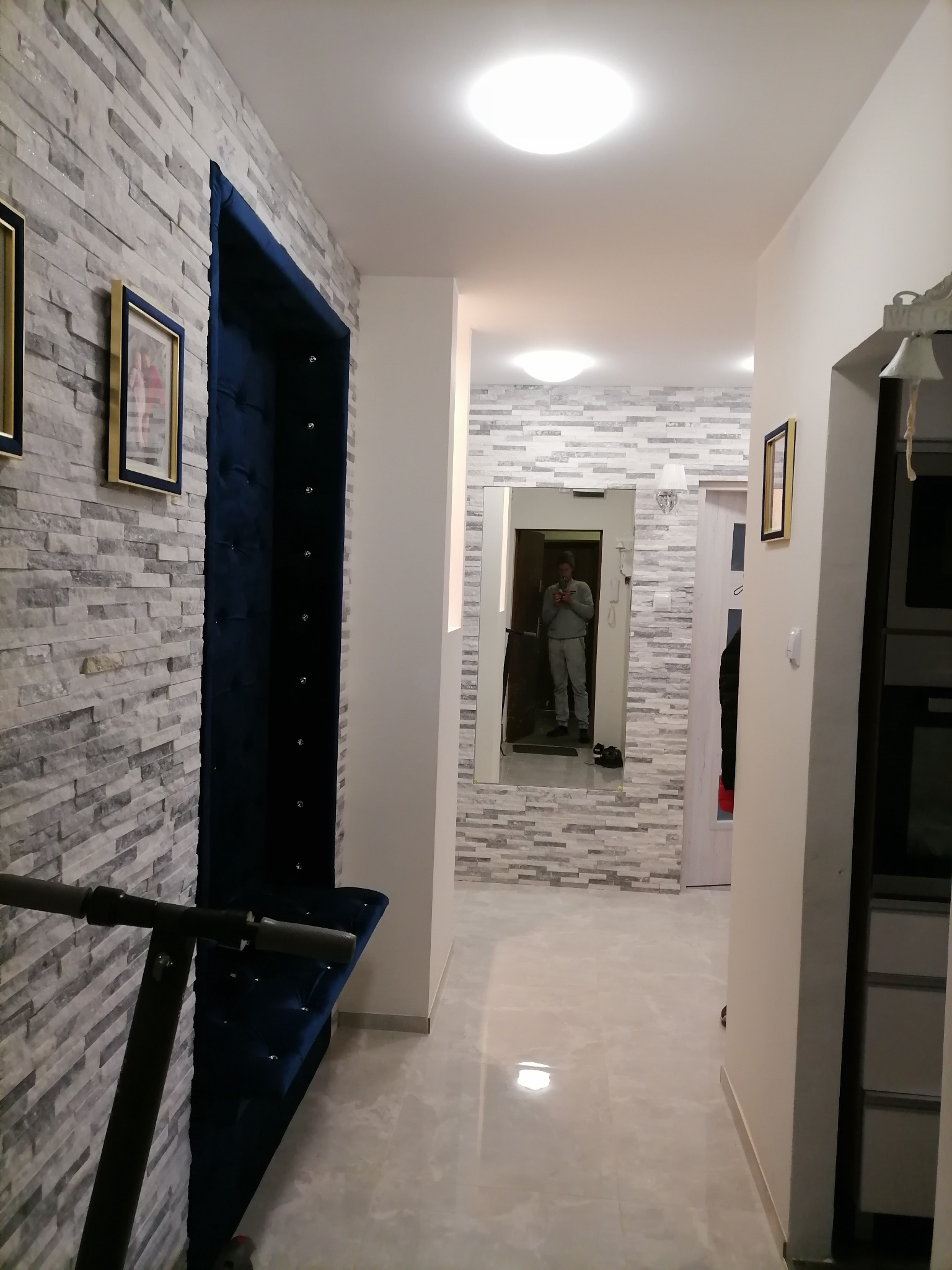 Koszalin: Korytarz z dekoracyjną ścianą z imitacji kamienia, obok wnęka z tapicerowanym na granatowo siedziskiem, lustro na końcu korytarza odbija wnętrze mieszkania.