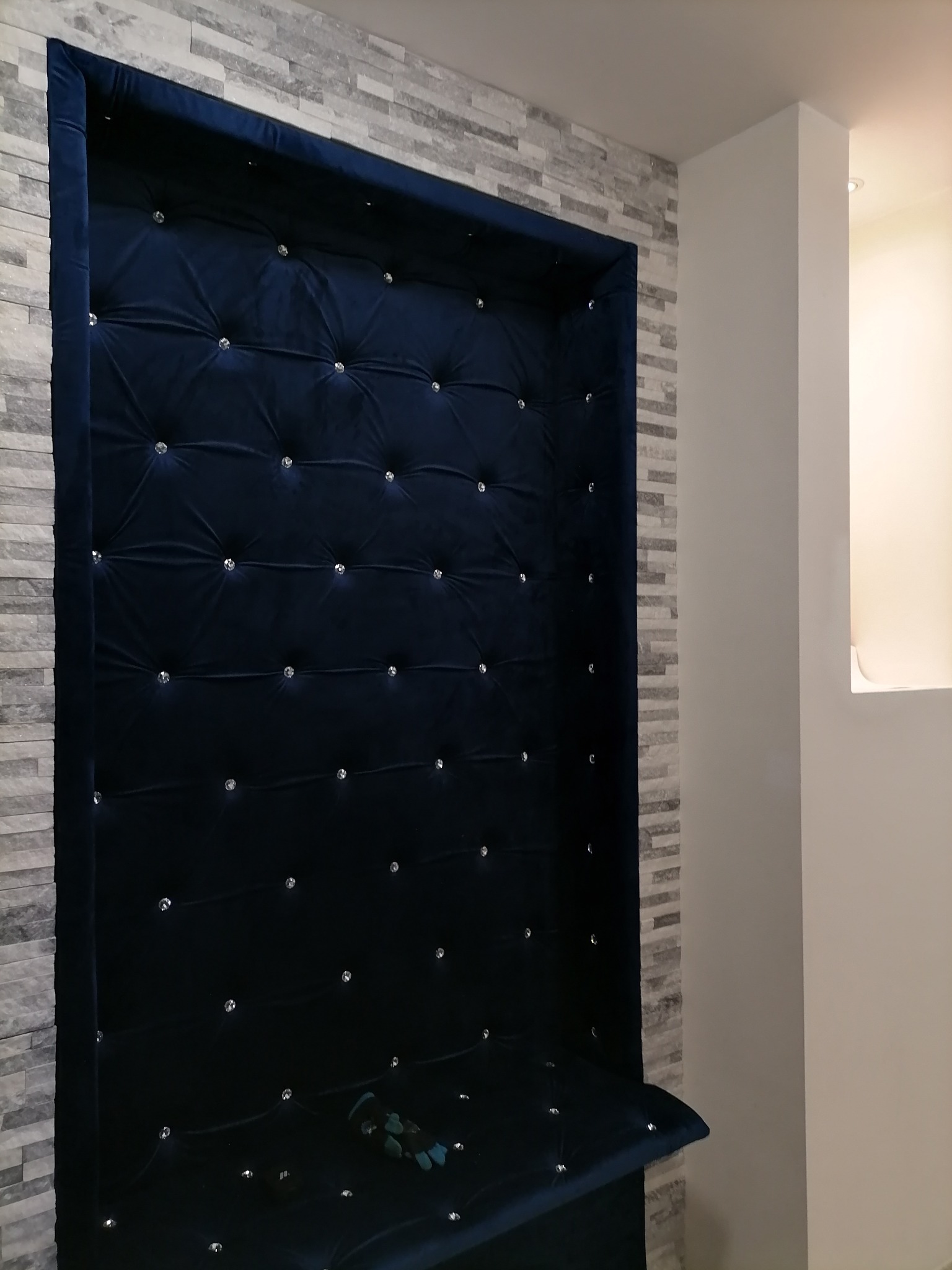 Niebieski, pikowany panel ścienny z kryształkami, wkomponowany między ścianę z dekoracyjnej cegły a białą ścianę z wnęką.