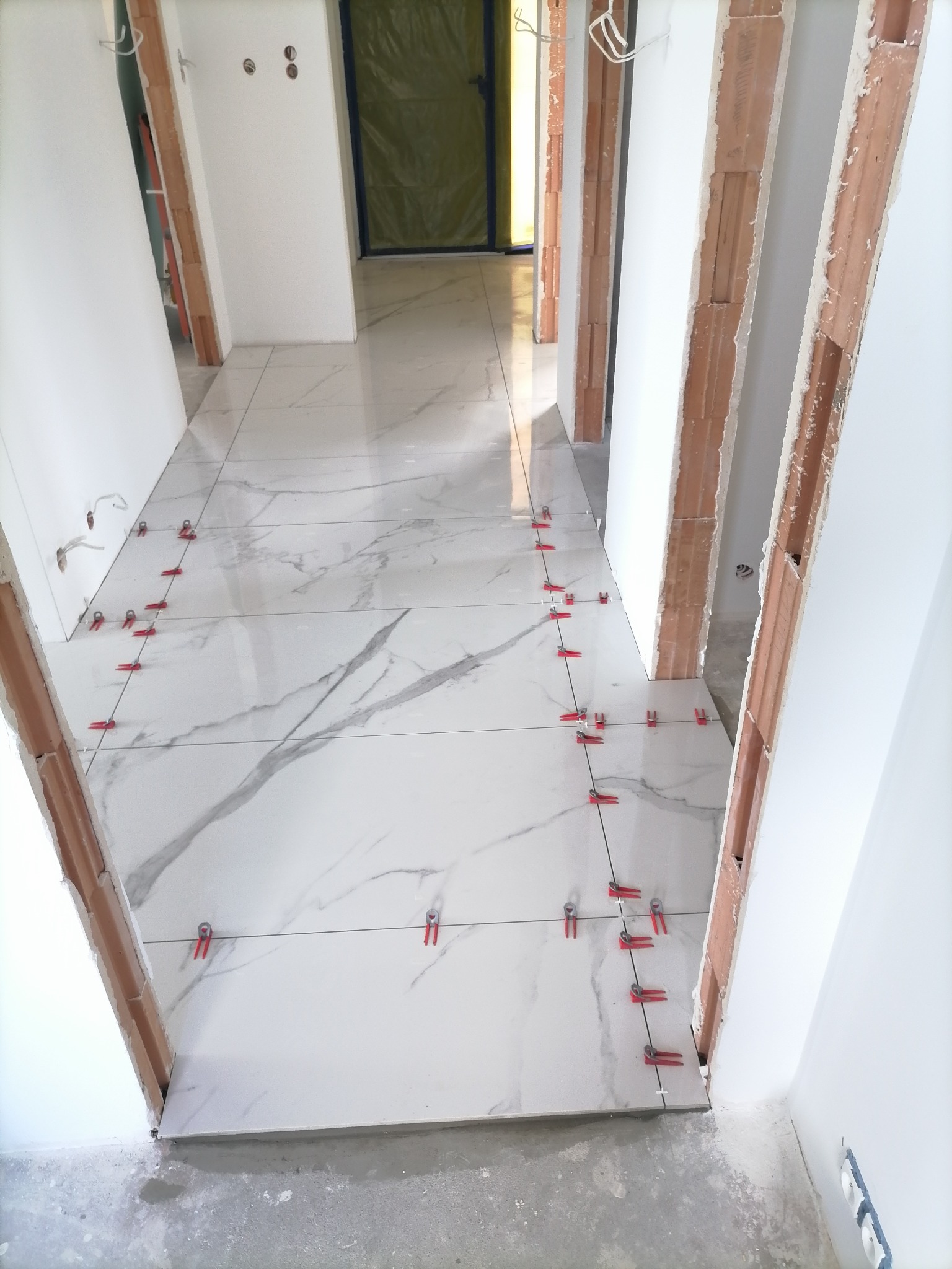 Układanie białych płytek podłogowych z szarym wzorem marmuru w korytarzu, widoczne czerwone klipsy poziomujące i niedokończone ościeżnice drzwi.