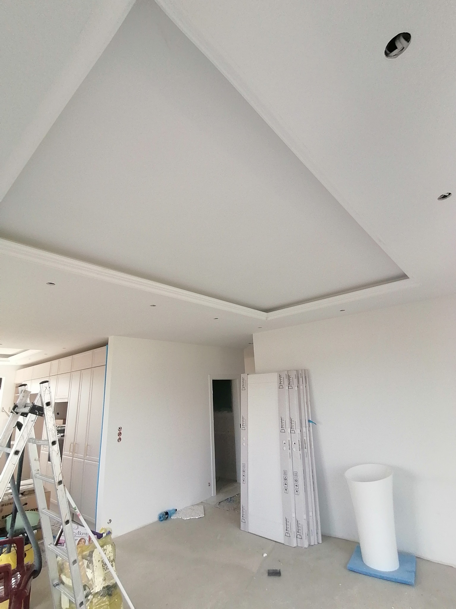 Biały sufit z podwieszanym fragmentem i dekoracyjną listwą, przygotowany pod montaż oświetlenia, widoczne otwory, w tle drabina i przygotowane panele ścienne.
