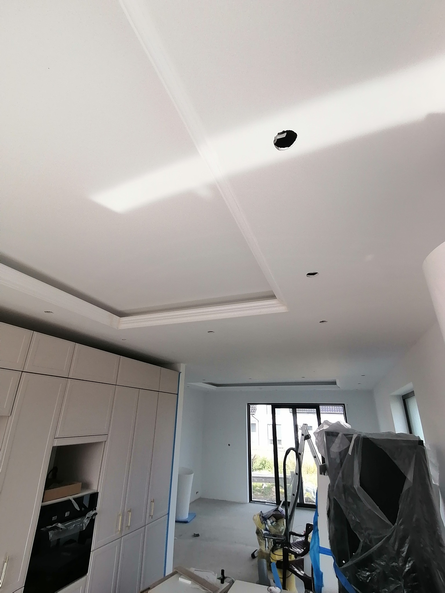 Biały sufit z dekoracyjnymi listwami i otworami na oświetlenie, widok z dołu, w tle zabudowa kuchenna i okno.