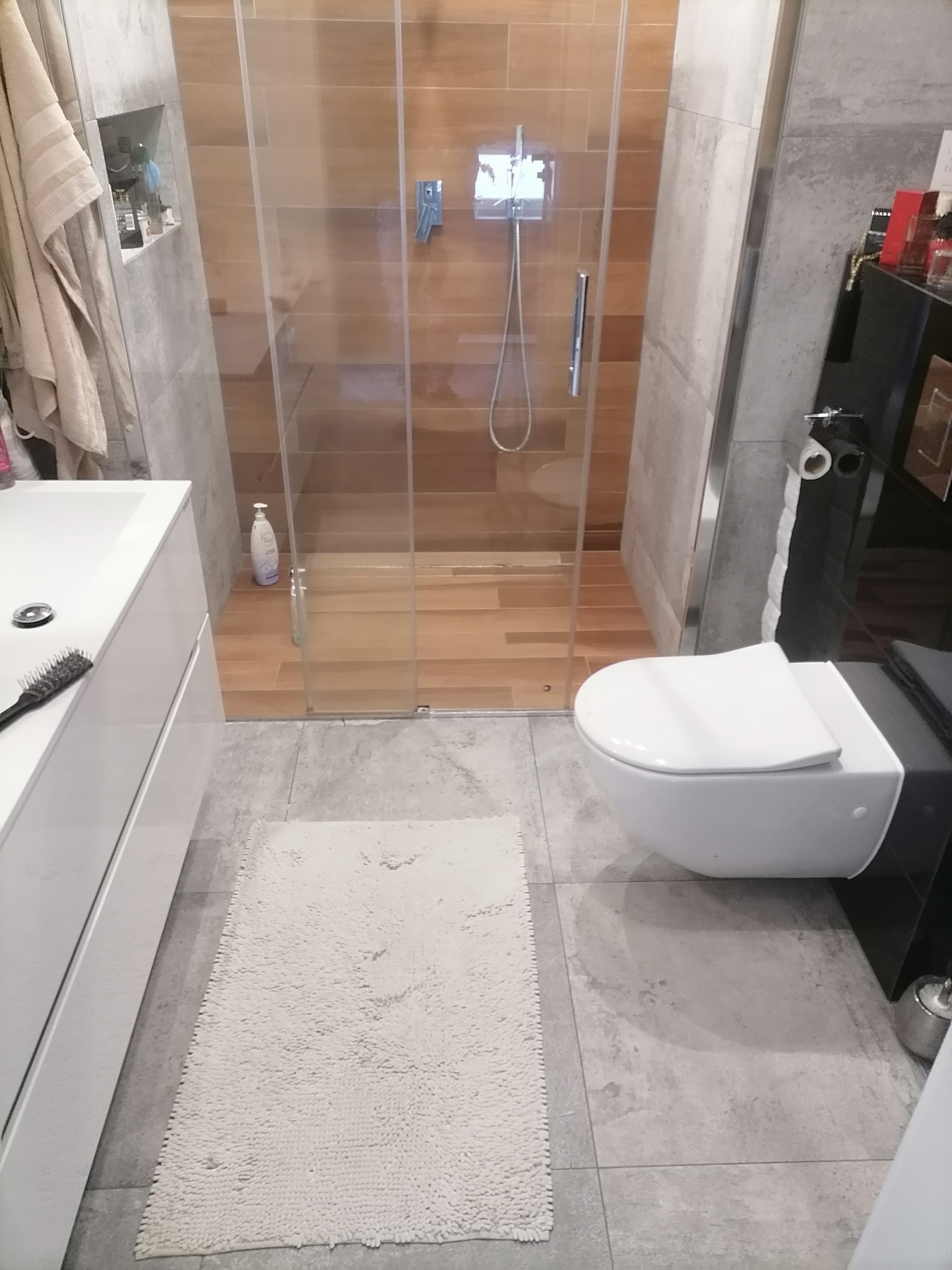 Nowoczesna łazienka z kabiną prysznicową typu walk-in, wiszącą toaletą i szarą płytką podłogową, uzupełniona o biały dywanik.