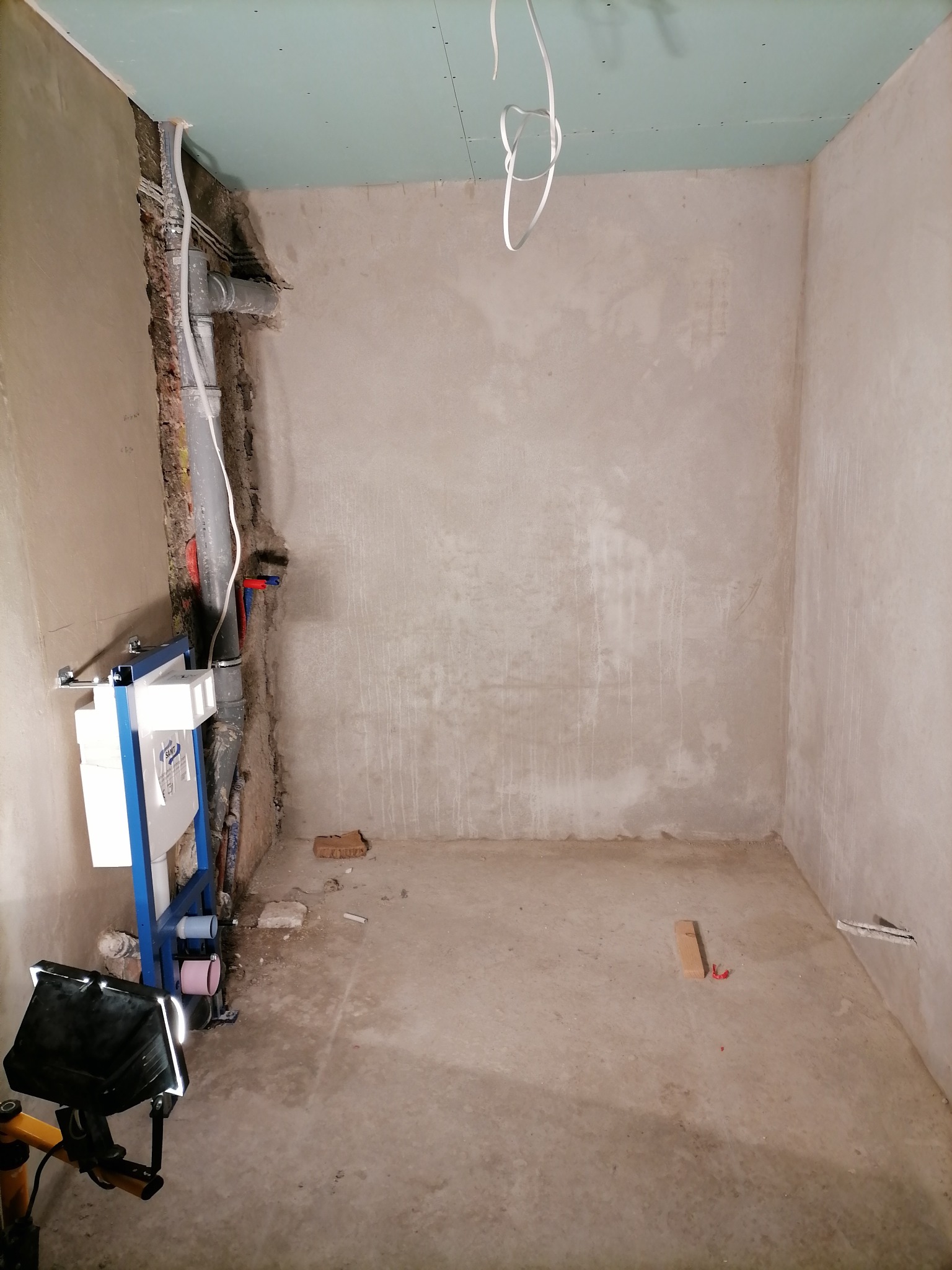 Wnętrze łazienki w trakcie remontu: surowe ściany, zamontowany stelaż podtynkowy WC z niebieską ramą, widoczne rury instalacyjne, częściowo położona płyta karton-gips na suficie, lampa budowlana...