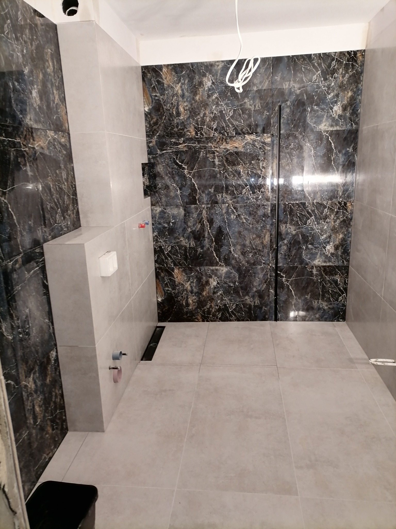 Wykończona łazienka z czarno-złotymi płytkami imitującymi marmur na ścianach, szare płytki na podłodze i ścianach, widoczne elementy instalacji sanitarnej i odpływ liniowy.