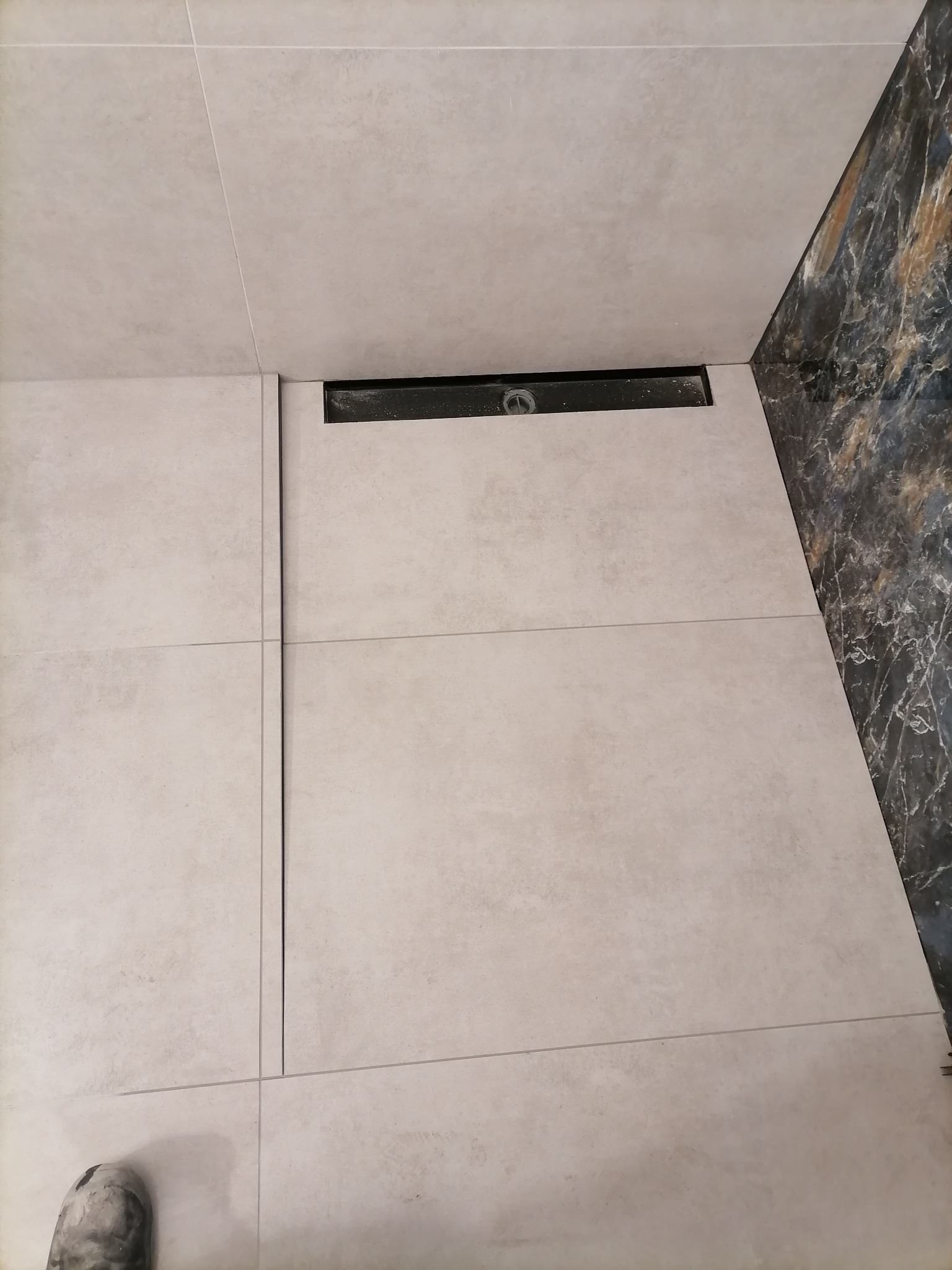 Jasne, kwadratowe płytki podłogowe z czarnym odpływem liniowym, widoczne fragmenty marmurowej ściany i but roboczy w rogu kadru.