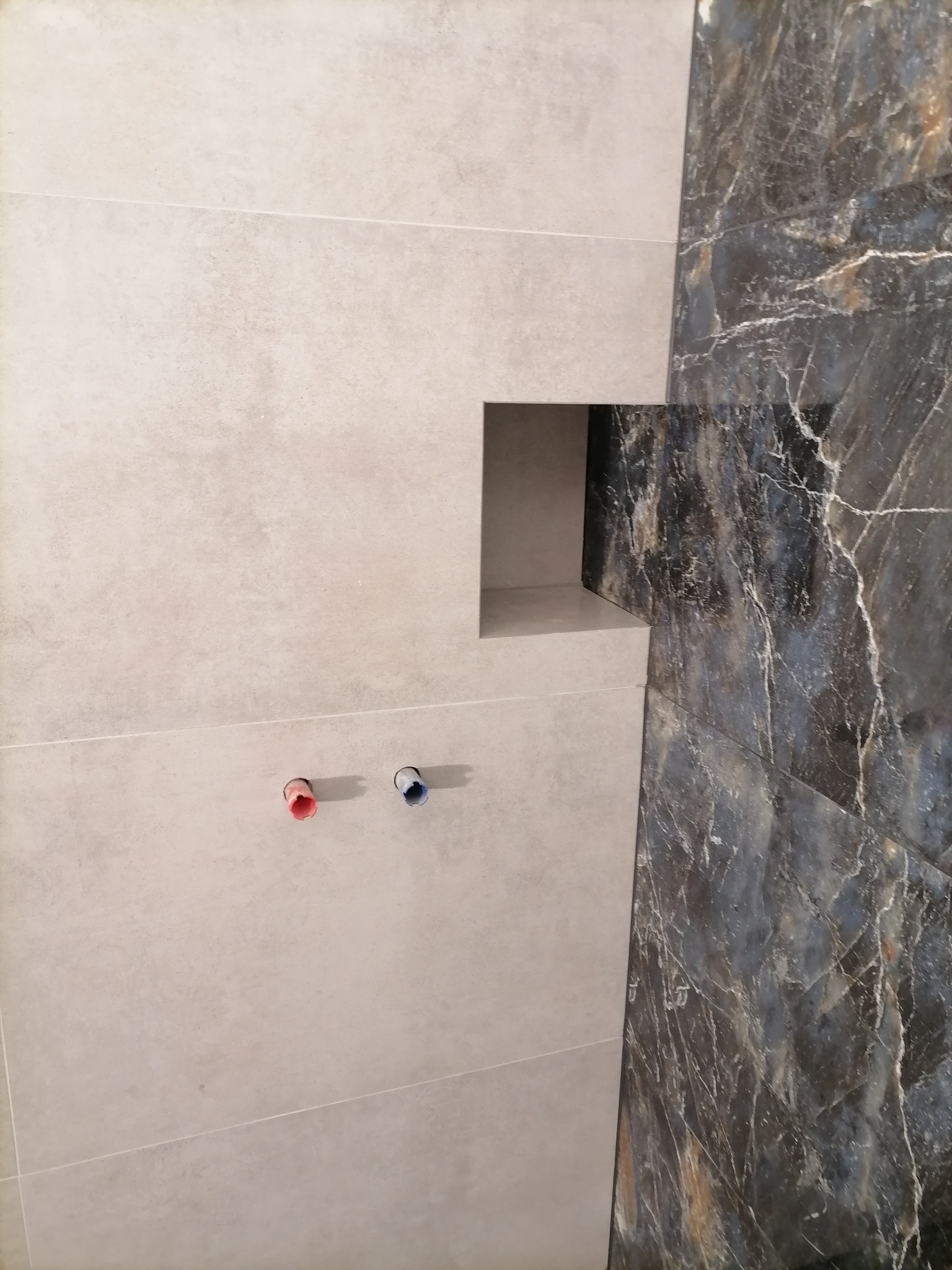 Ściana łazienki wyłożona jasnymi, matowymi płytkami z wnęką i widocznymi rurami z czerwonym i niebieskim oznaczeniem, sąsiadująca z ścianą z ciemnych płytek imitujących marmur z wyraźnym żyłowaniem.