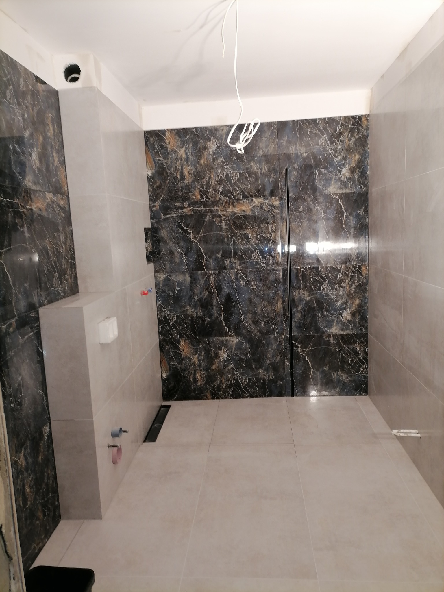 Wnętrze łazienki w trakcie prac wykończeniowych, widoczne płytki imitujące marmur na ścianie, jasne płytki podłogowe i częściowo zabudowany stelaż podtynkowy WC z wystającymi rurami.