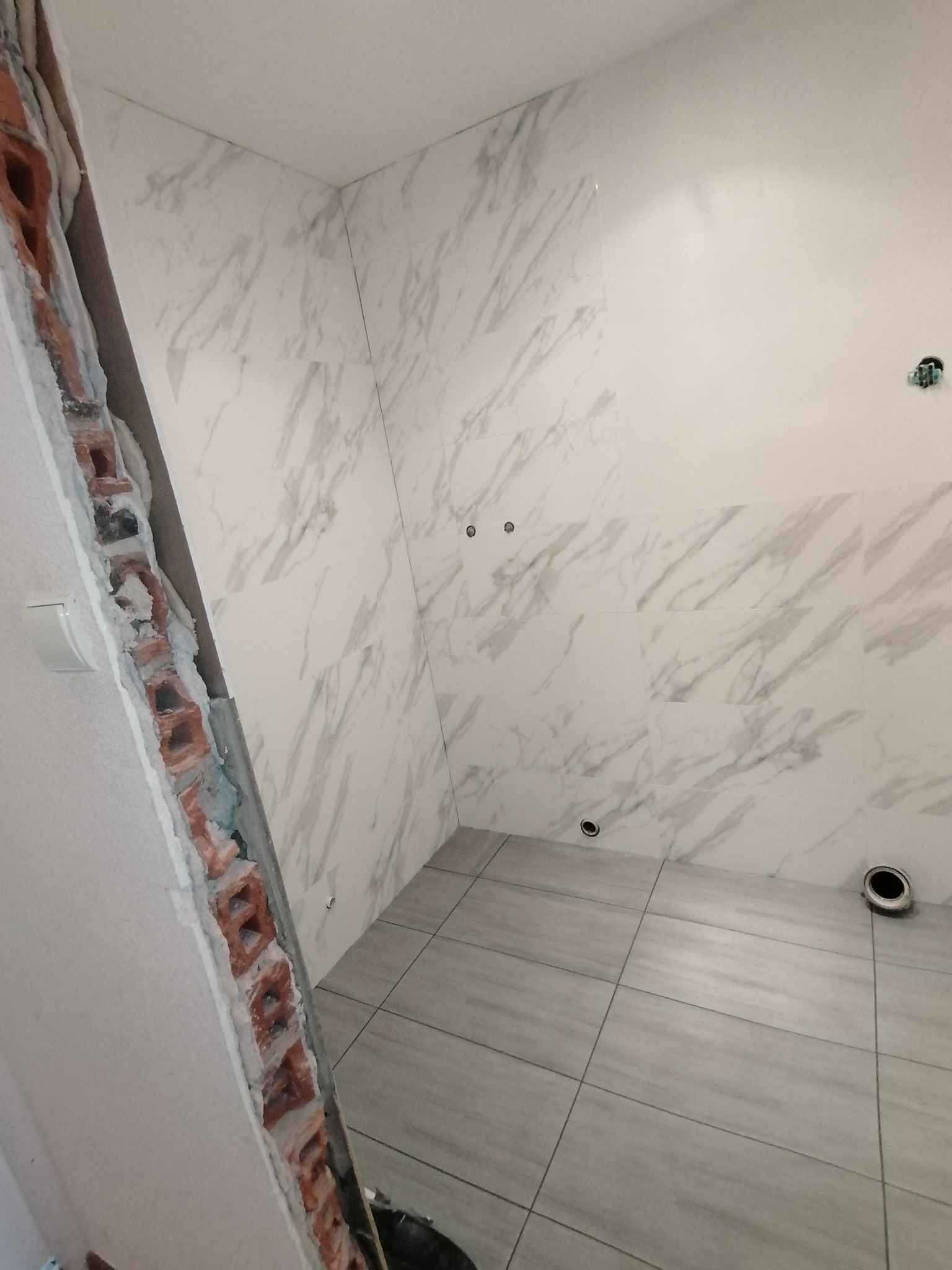 Wnętrze łazienki w trakcie remontu, widoczne płytki imitujące marmur na ścianach i podłodze, odsłonięta cegła przy ścianie, przygotowane otwory na armaturę.