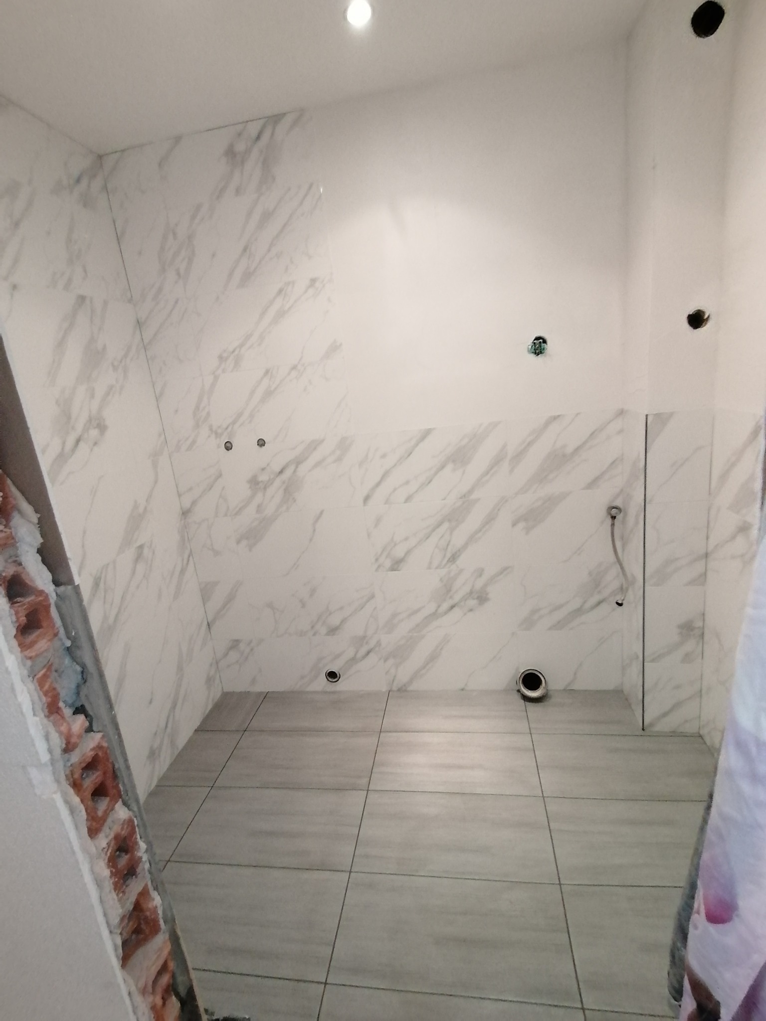 Łazienka w trakcie remontu: ściany częściowo wyłożone płytkami imitującymi marmur, widoczne otwory na instalacje, surowy mur w rogu pomieszczenia, szare płytki podłogowe.