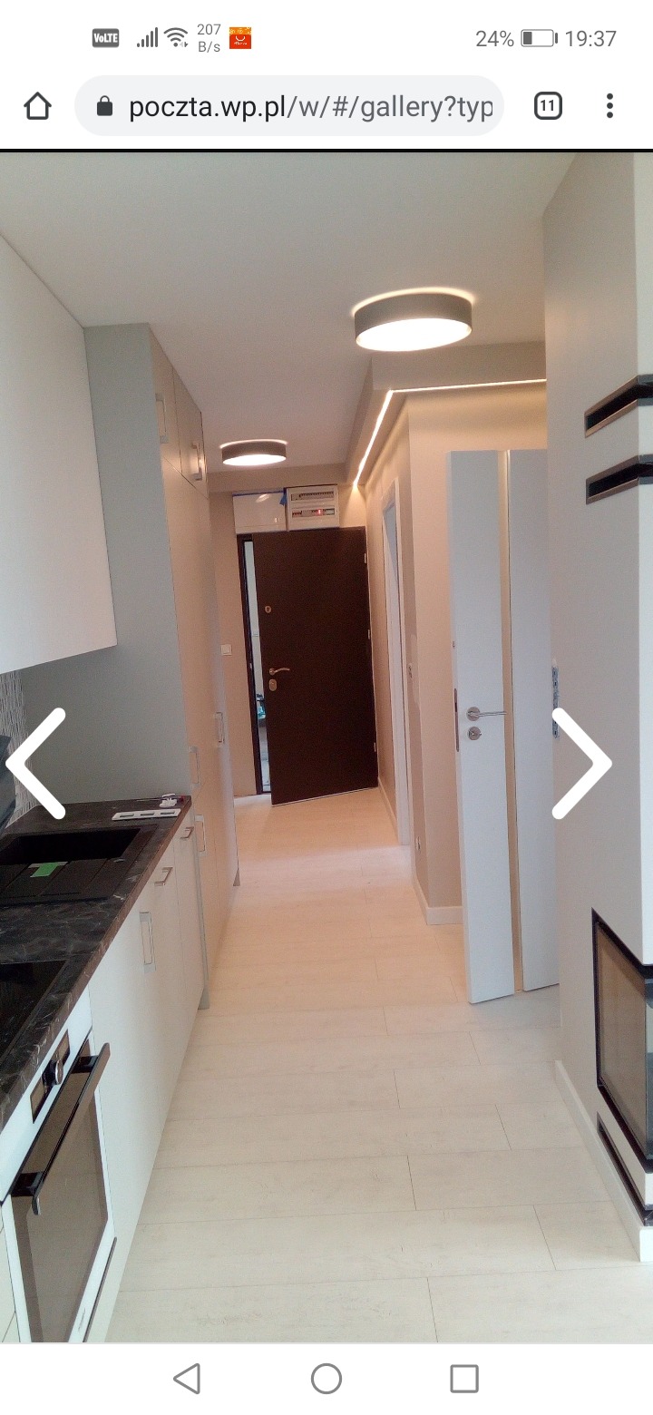 Nowoczesne wnętrze mieszkania z jasną podłogą imitującą drewno, zabudową kuchenną w odcieniach bieli i szarości oraz minimalistycznym oświetleniem sufitowym LED.