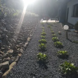 Świeżo założony ogród z rabatą z kamieni, wysypany drobnym szarym żwirem, z rzędem młodych roślin i dwoma kulistymi lampami ogrodowymi, oświetlony silnym słońcem.