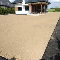 Przygotowany teren pod trawnik przy nowoczesnym domu z drewnianymi elementami, widoczne obrzeża trawnikowe i tuje wzdłuż ogrodzenia.