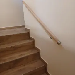 Schody wyłożone panelami imitującymi drewno, z jasną drewnianą poręczą mocowaną do ściany, wykończone listwami przypodłogowymi w kolorze paneli.